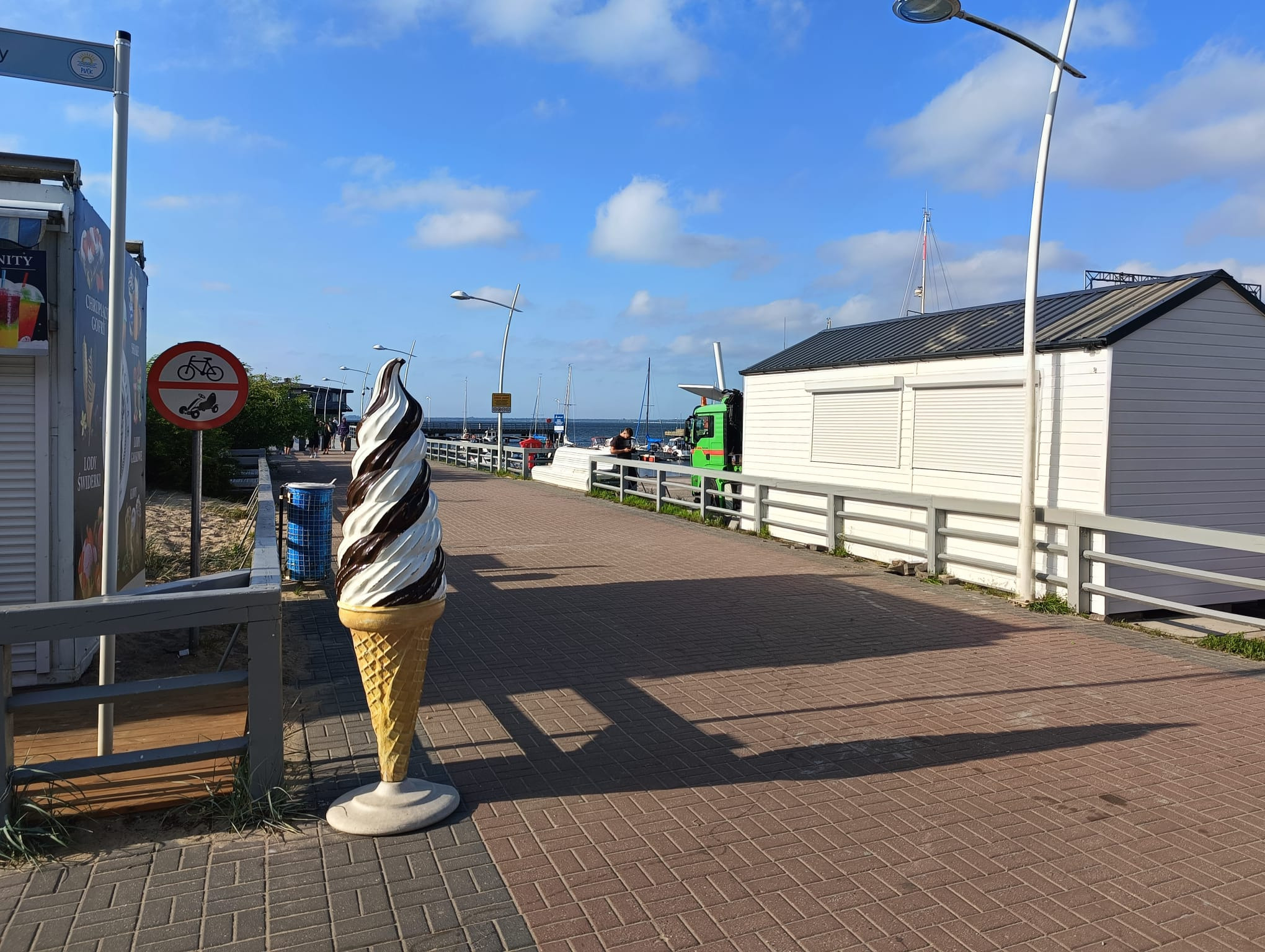Duża rzeźba lodów w rożku, z białymi i brązowymi spiralami, stoi na brukowanej promenadzie w słoneczny dzień, w tle budki handlowe i błękitne niebo.