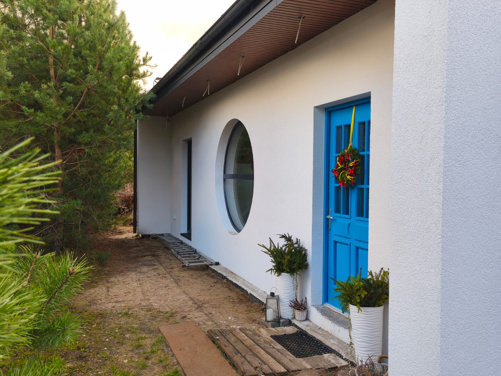 Biały budynek z okrągłym oknem i niebieskimi drzwiami ozdobionymi wieńcem, obok rosną drzewa iglaste, na ziemi leży drabina.