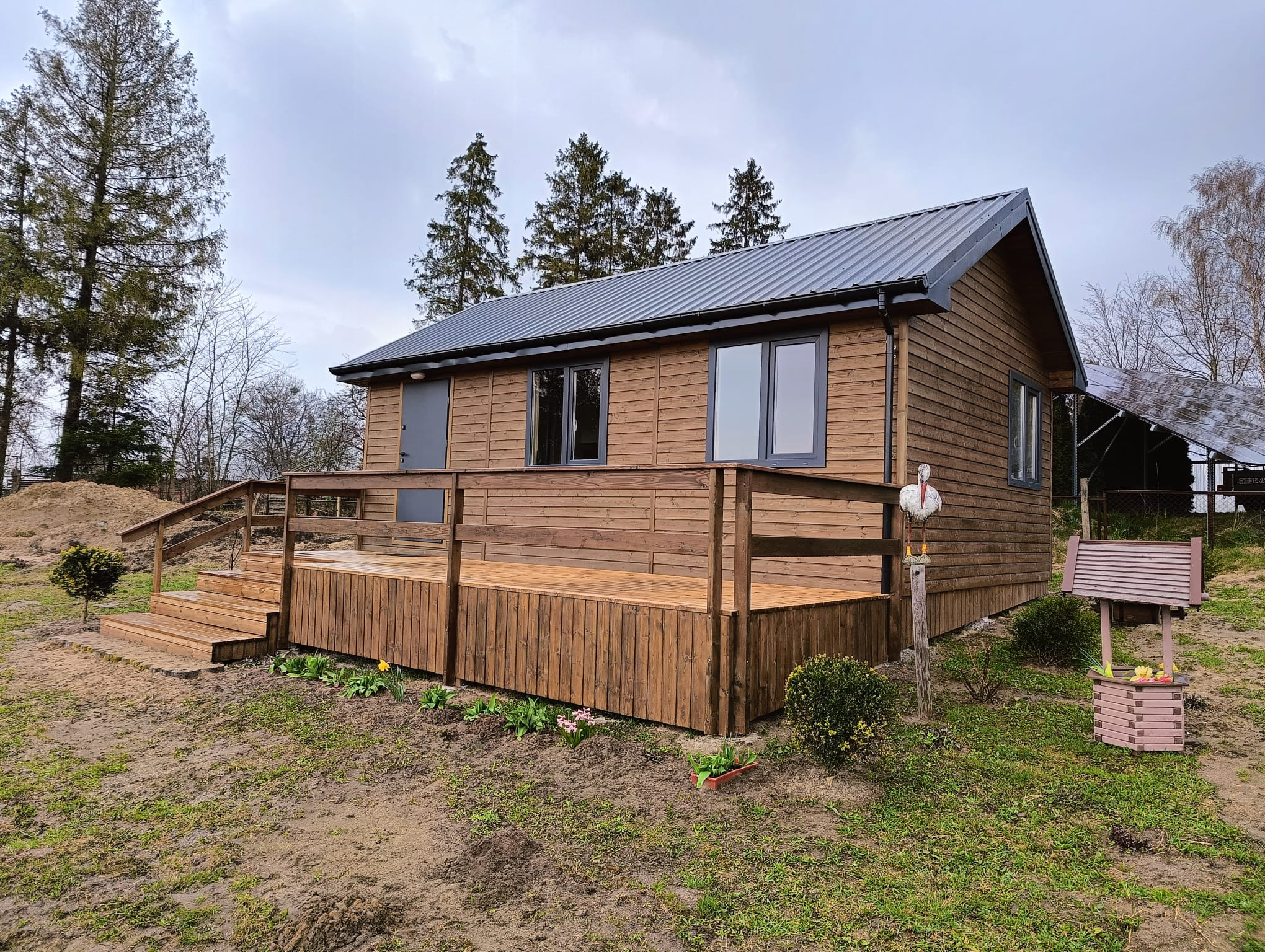 Niewielki, parterowy dom z drewnianą elewacją i metalowym dachem, z przyległym tarasem z drewnianą balustradą, ozdobiony figurką bociana i dekoracyjną studnią.