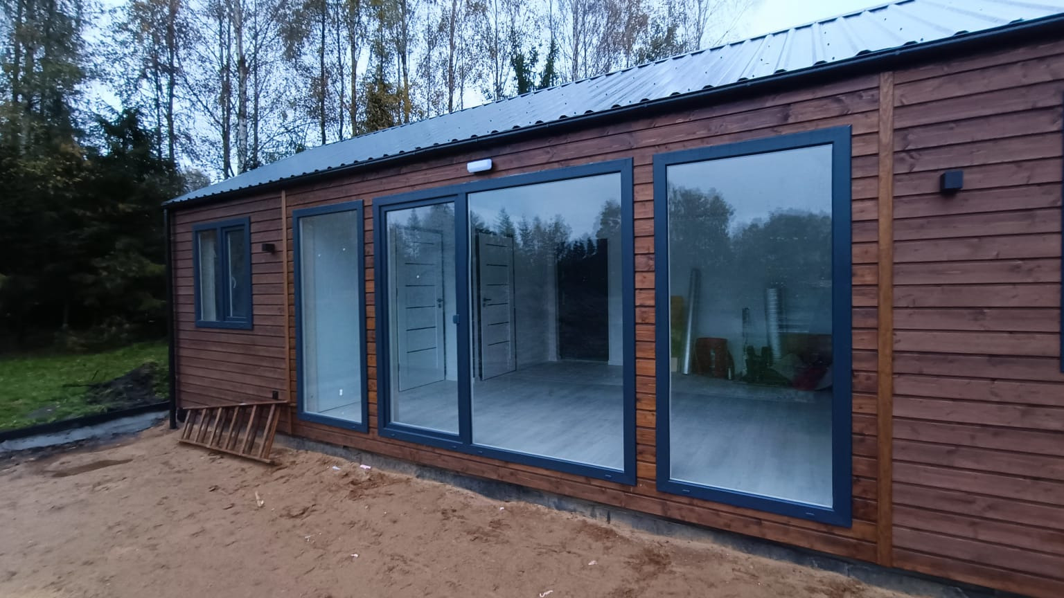 Nowoczesny, parterowy budynek z drewnianą elewacją, dużymi oknami i metalowym dachem na działce z piaskiem. Wnętrze widoczne przez okna, z szarymi panelami podłogowymi i białymi drzwiami.