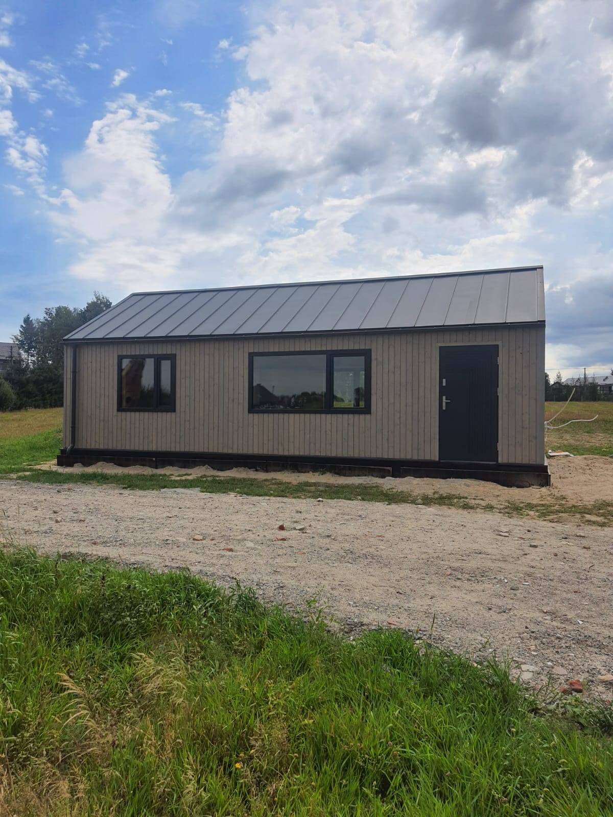 Gotowy, parterowy dom szkieletowy z szarymi panelami elewacyjnymi, czarną stolarką okienną i drzwiową, oraz metalowym dachem na działce z gruntem i zielenią.