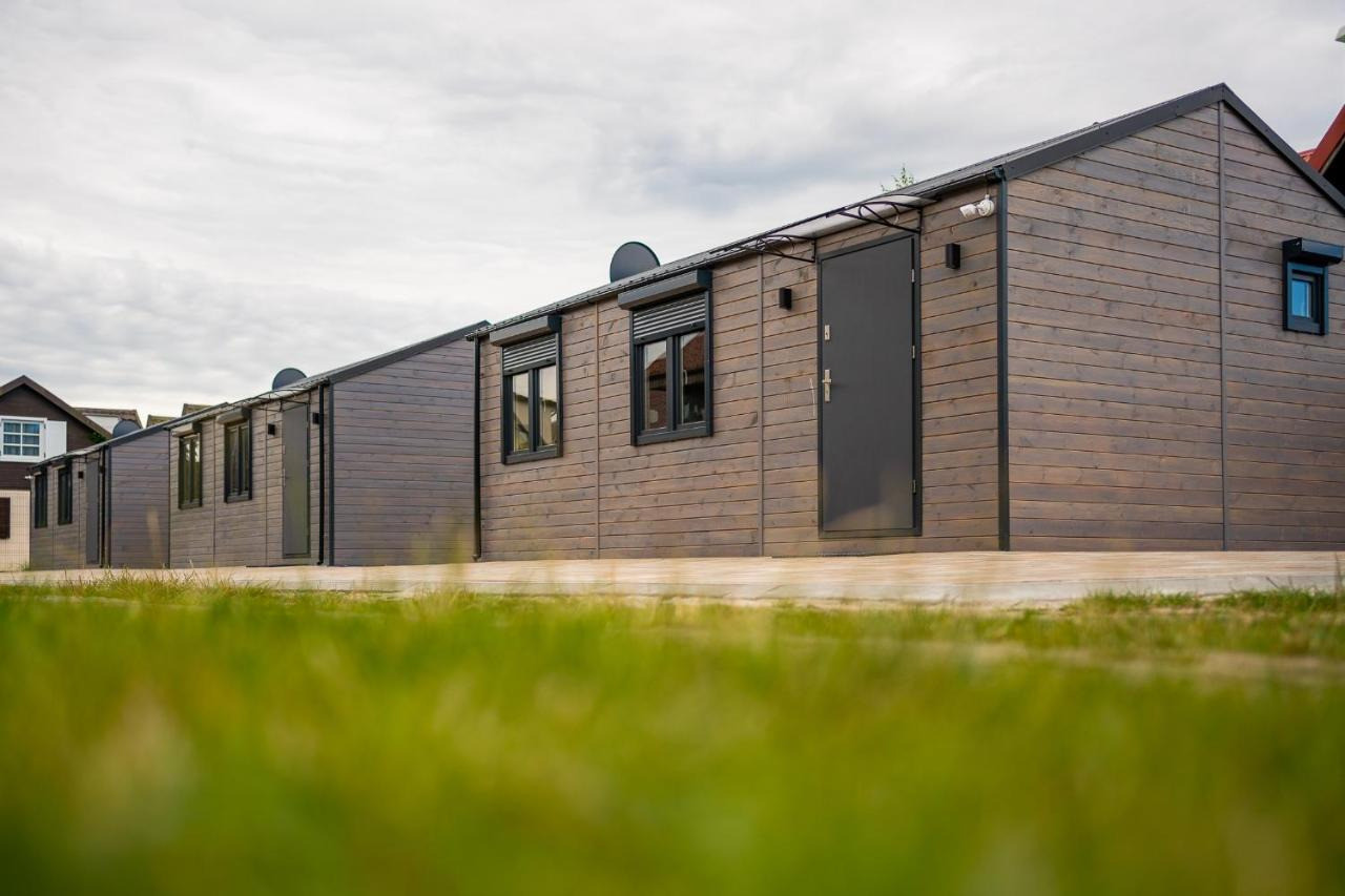 Szereg modułowych budynków z drewnianą elewacją, z szarymi drzwiami i oknami z roletami zewnętrznymi, widziane z perspektywy trawnika.