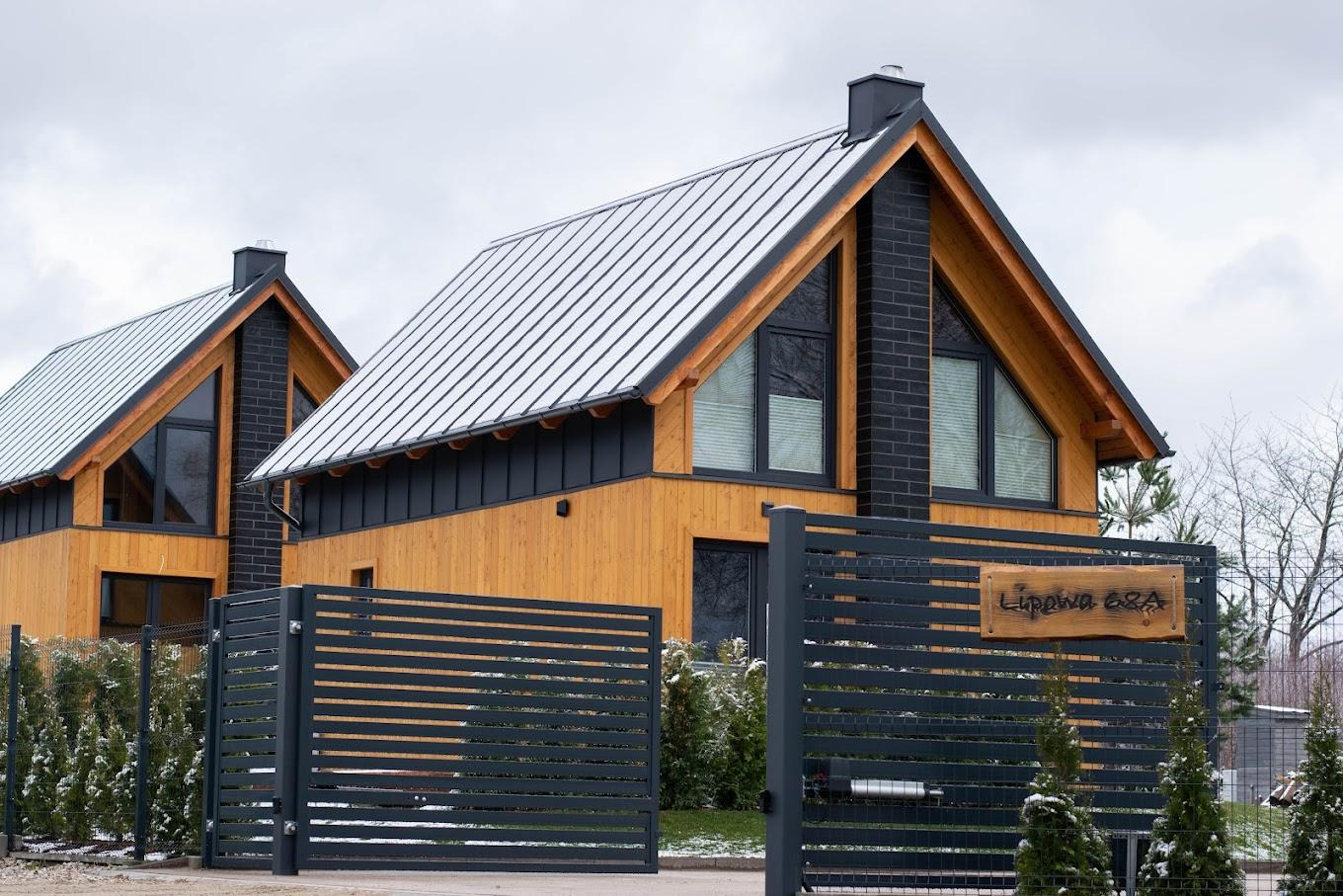 Nowoczesny dom z drewnianą elewacją, czarnym kominem z cegły i metalowym dachem, widoczny przez otwartą, poziomą bramę w kolorze antracytowym z drewnianą tabliczką adresową.