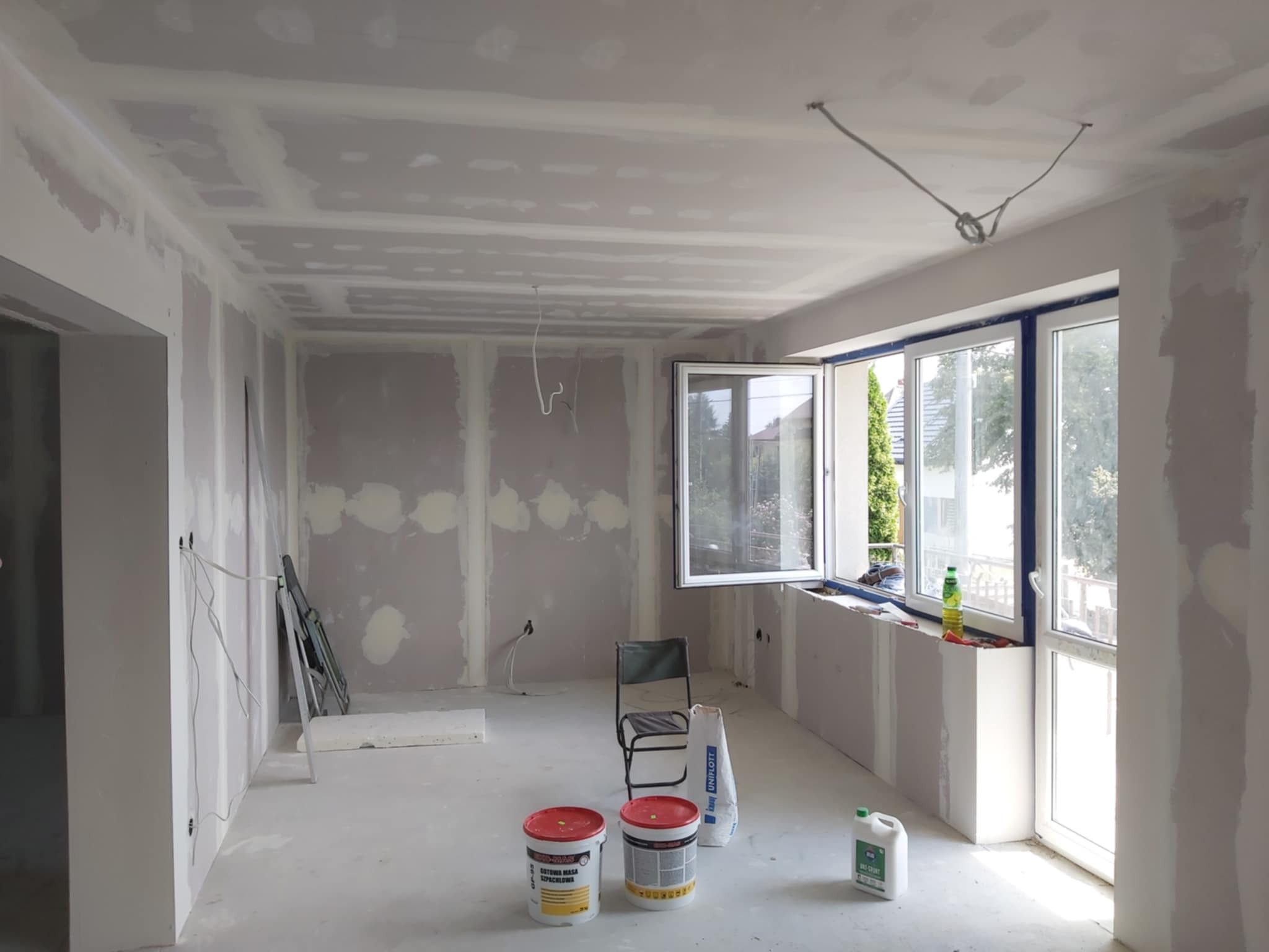 Pokój w trakcie remontu, ściany i sufit pokryte płytami kartonowo-gipsowymi przed szpachlowaniem, widoczne puszki z masą szpachlową, krzesło i worki z materiałami budowlanymi.