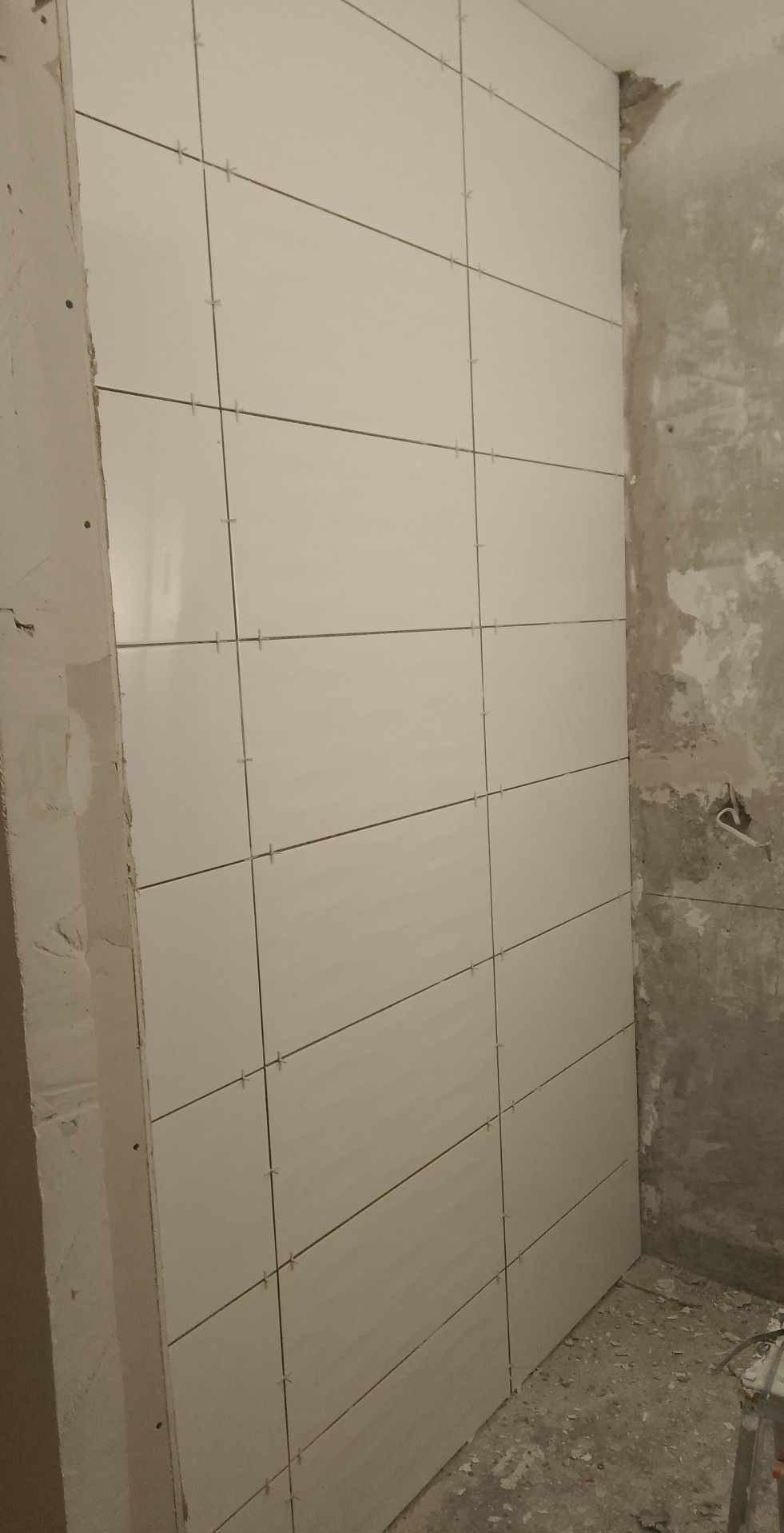 Świeżo ułożone, jasne płytki ceramiczne na ścianie w trakcie remontu, z widocznymi krzyżykami dystansowymi i surową ścianą obok.