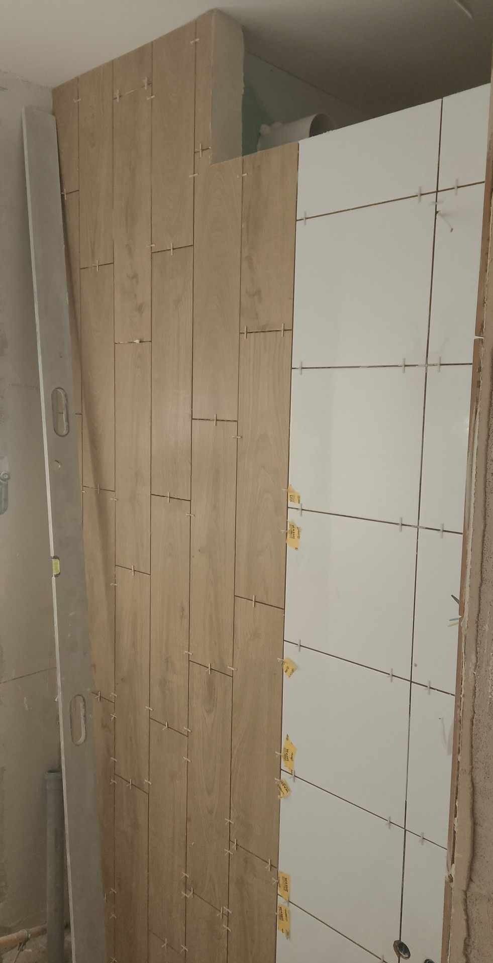 W trakcie układania płytek imitujących drewno na ścianie, obok białe płytki z krzyżykami dystansowymi, poziomica oparta o ścianę.