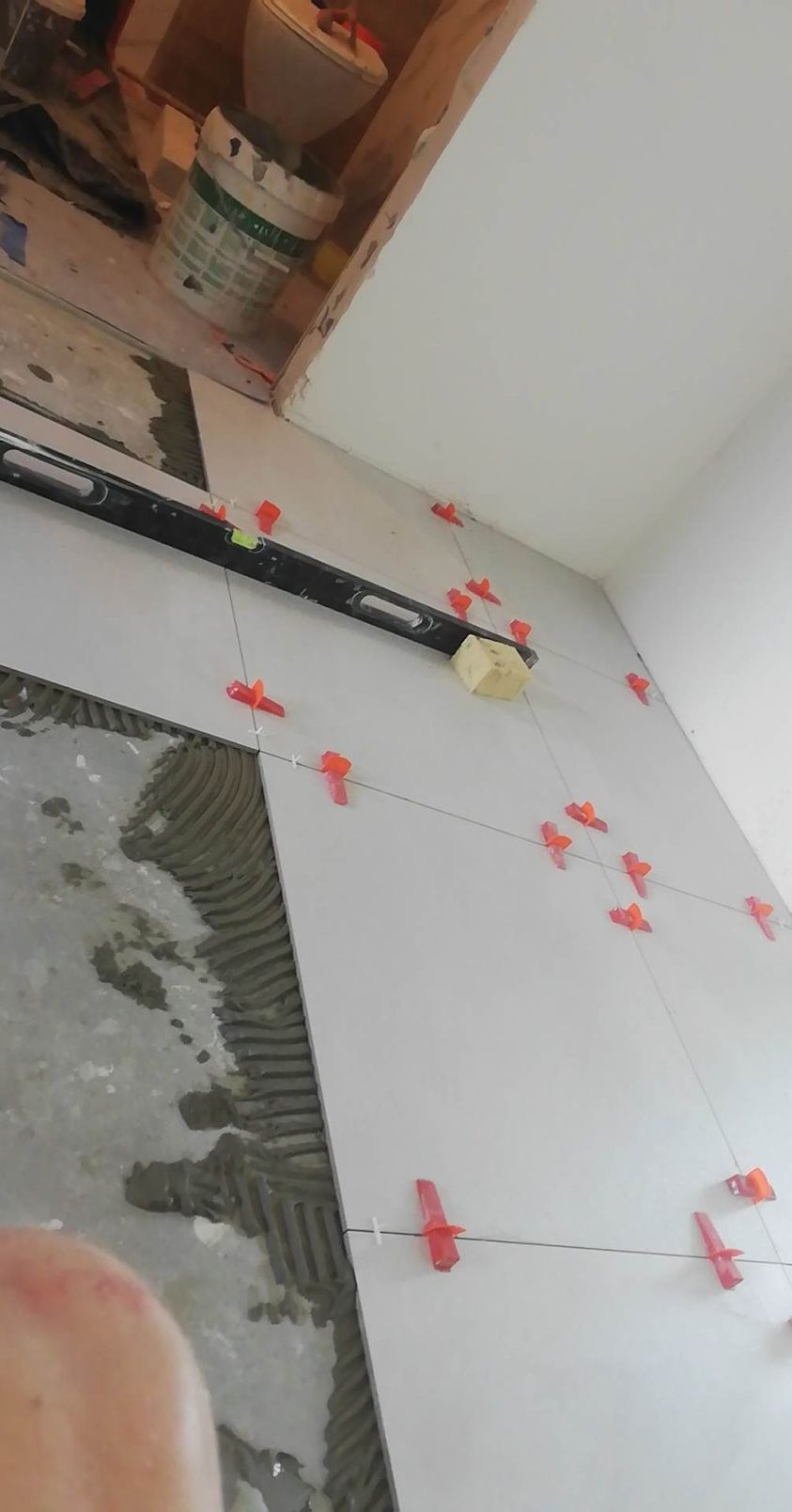 Układanie białych płytek na ścianie z użyciem poziomicy, sznurka i czerwonych krzyżyków dystansowych, widoczna warstwa kleju na fragmencie ściany.