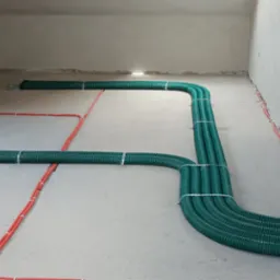 Szczegółowy widok podłogi w surowym stanie z rozłożonymi rurami wentylacyjnymi w kolorze zielonym i przewodami elektrycznymi w kolorze pomarańczowym, zamocowanymi do podłoża przy użyciu metalowych...