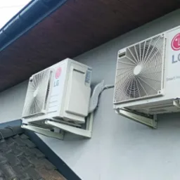 Dwa klimatyzatory LG Smart Inverter zamontowane na elewacji budynku z widocznymi wspornikami i instalacją odprowadzającą skropliny, z fragmentem dachu i rynny w tle.