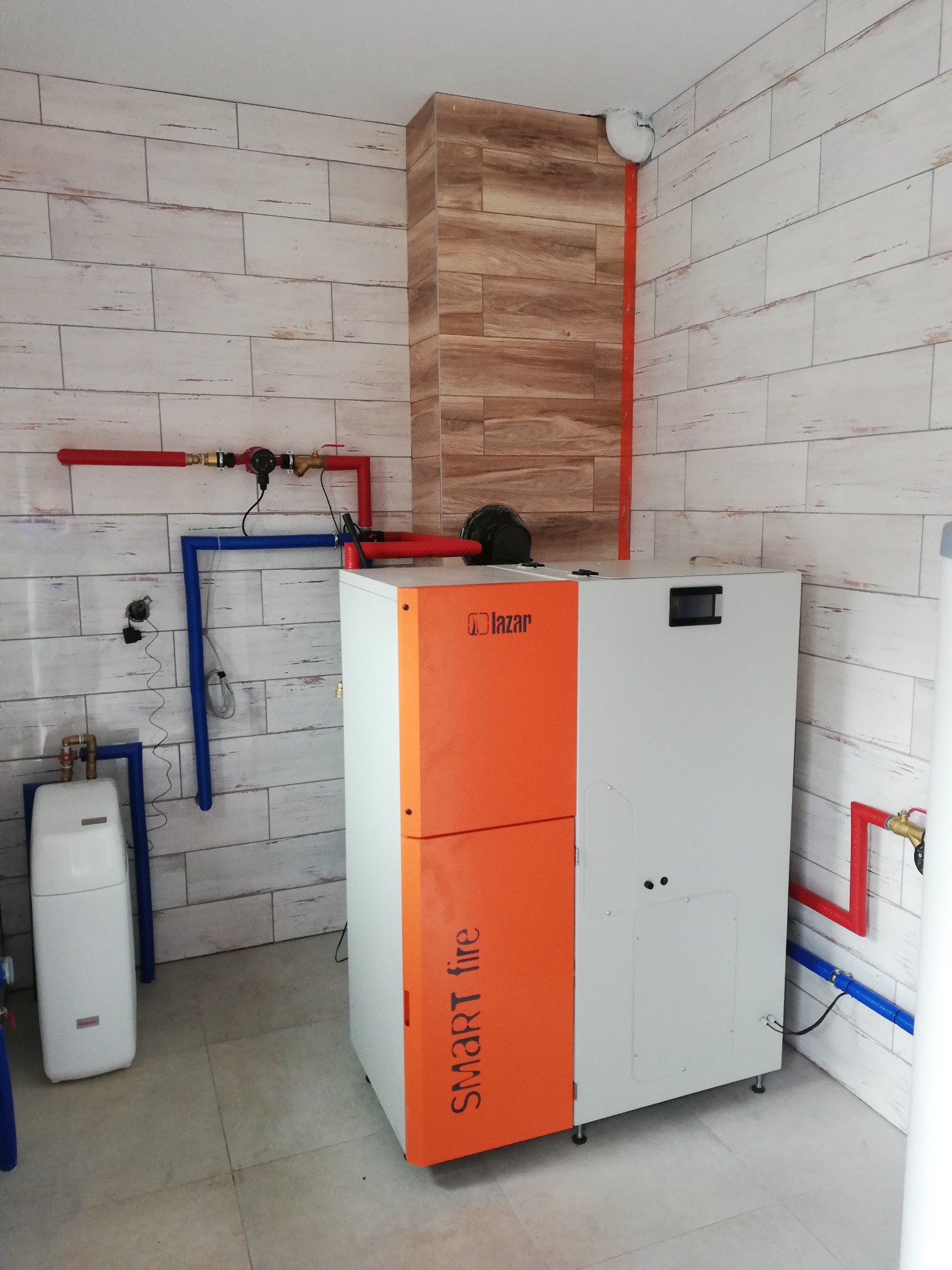 Nowoczesny piec CO marki Lazar Smart Fire w kotłowni wykończonej płytkami imitującymi drewno, z widocznymi elementami instalacji grzewczej: rurami w kolorze czerwonym i niebieskim, oraz zasobnikiem...