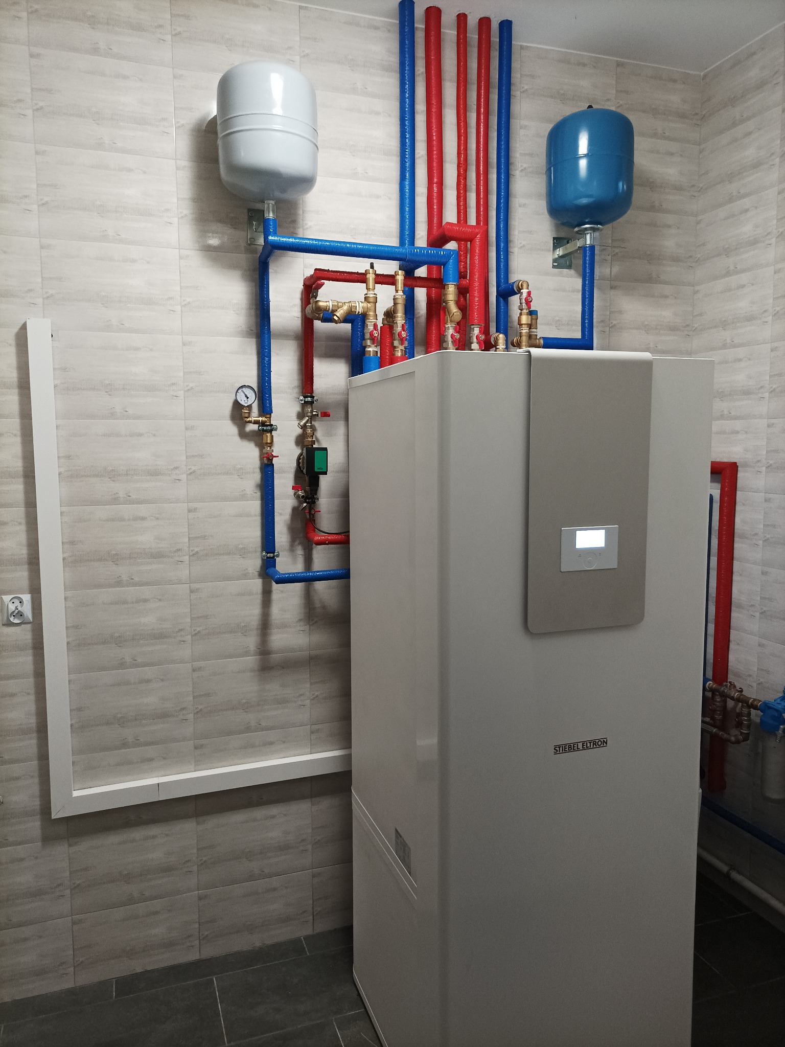 Nowoczesny, biały piec centralnego ogrzewania marki Stiebel Eltron z podłączonymi rurami w kolorach niebieskim i czerwonym oraz dwoma zbiornikami wyrównawczymi, zainstalowany w pomieszczeniu...