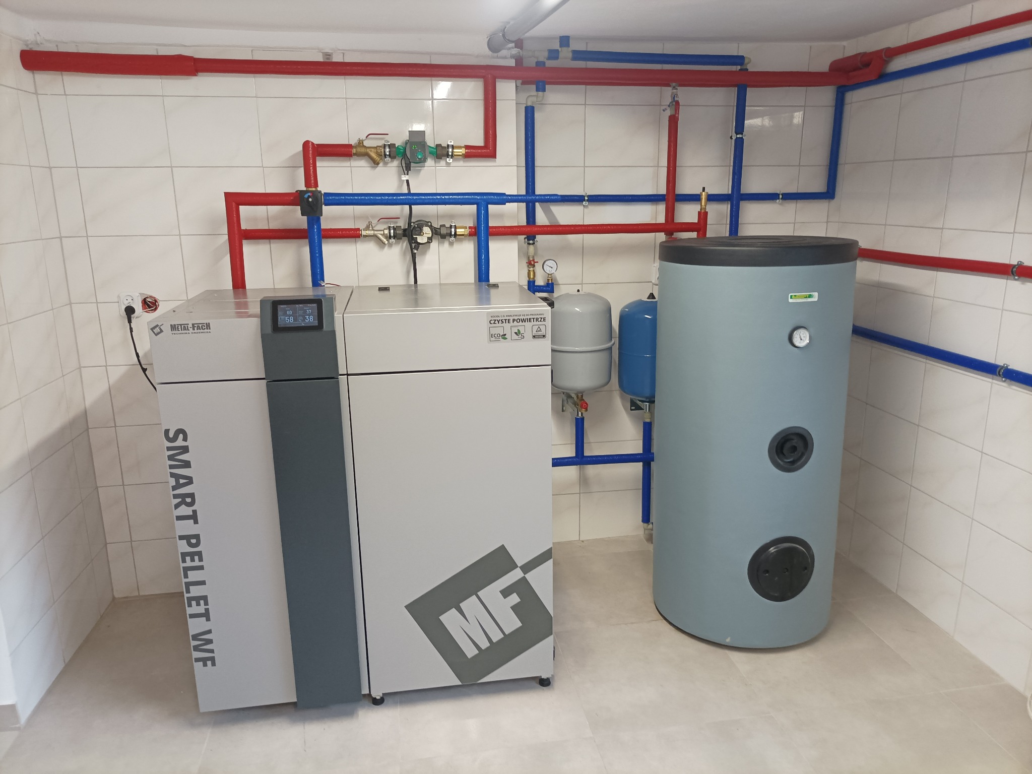 Nowoczesny piec na pellet z zasobnikiem wody, widoczne rury instalacji grzewczej w kolorach czerwonym i niebieskim, zamontowane w pomieszczeniu z białymi płytkami.