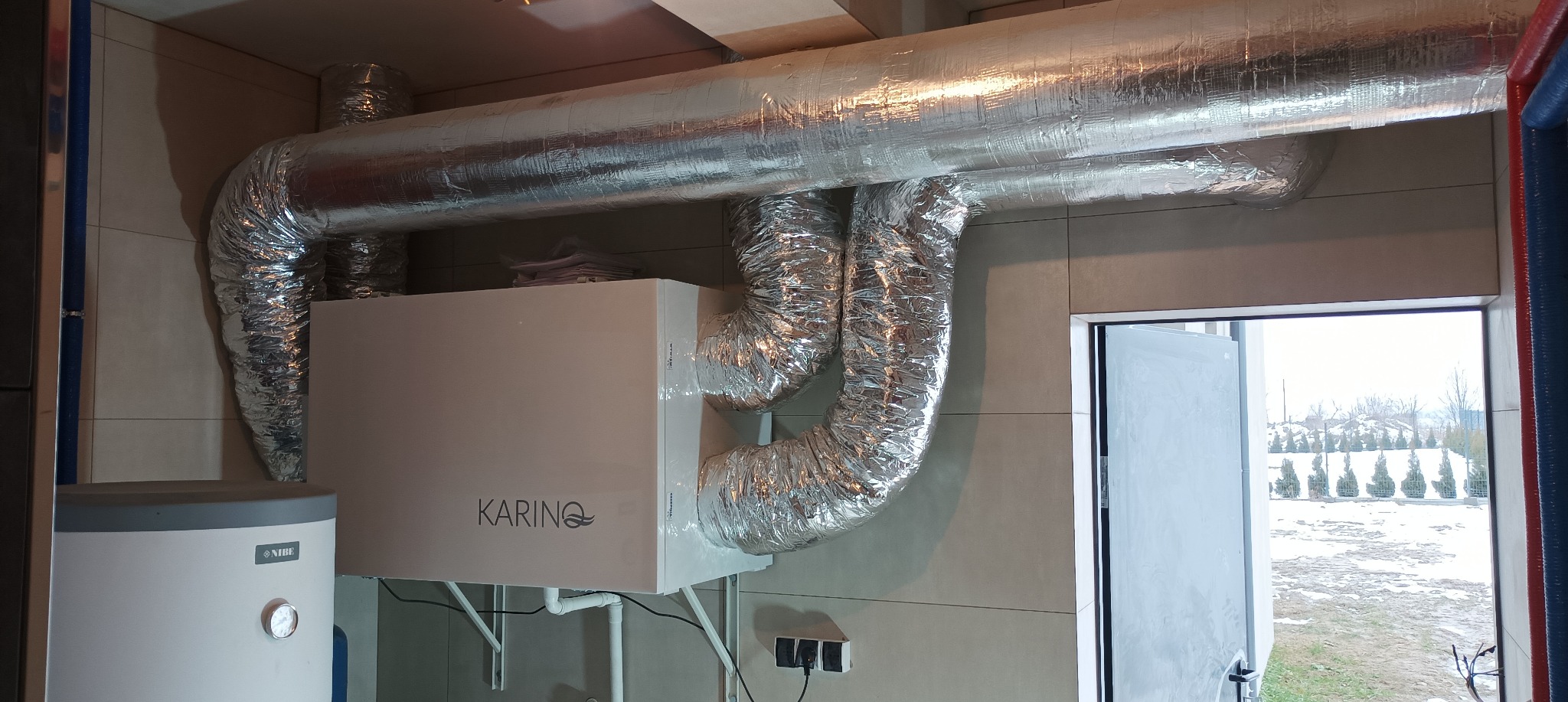 Centrala wentylacyjna z rekuperacją marki Karina, widoczne aluminiowe przewody wentylacyjne, bojler Nibe, oraz widok na zimowy krajobraz za oknem.