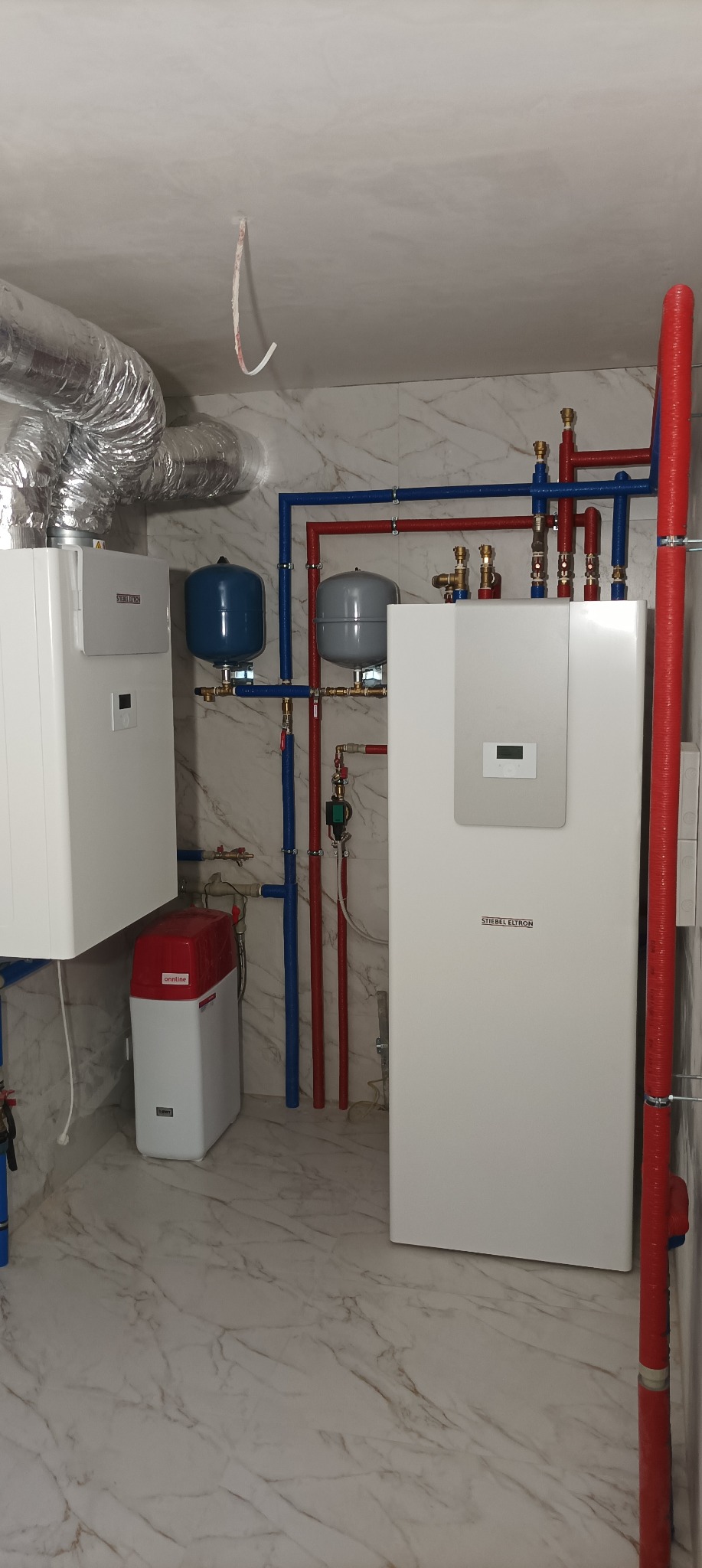 Kompletna instalacja urządzeń grzewczych Stiebel Eltron, z widocznymi zbiornikami wyrównawczymi, rurami w izolacji oraz zmiękczaczem wody, na tle marmurowej posadzki i ściany.