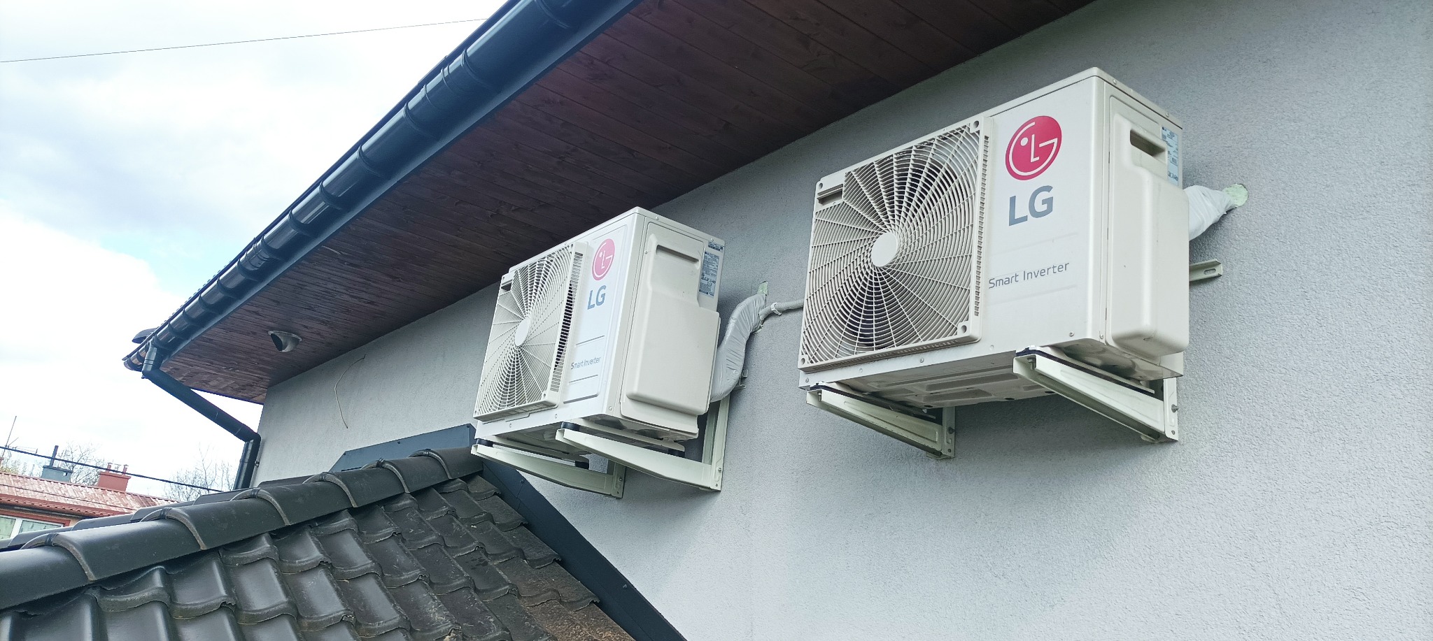 Dwa klimatyzatory LG Smart Inverter zamontowane na elewacji budynku z widocznymi wspornikami i instalacją odprowadzającą skropliny, z fragmentem dachu i rynny w tle.