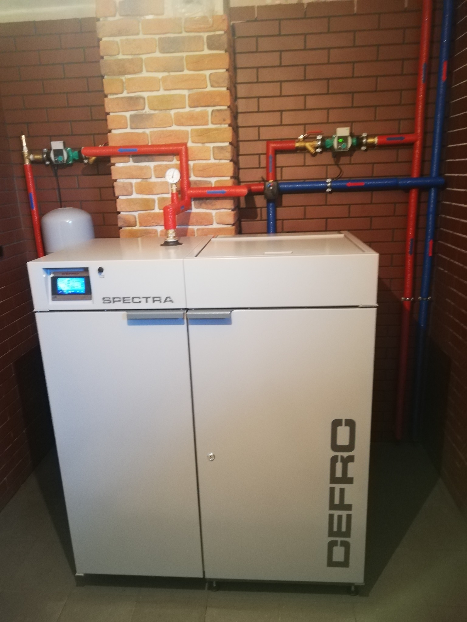 Nowoczesny piec CO marki DEFRO Spectra z widoczną instalacją hydrauliczną na tle ściany z cegły.