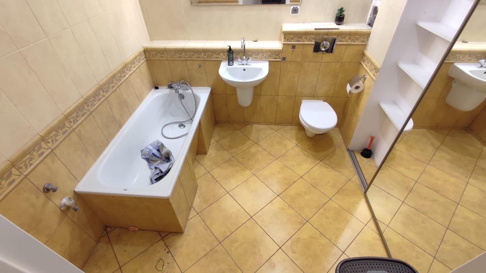 Ukończona łazienka z beżowymi płytkami, białą wanną z ręcznikiem w środku, umywalką i toaletą. Lustro z półkami odbija część pomieszczenia.