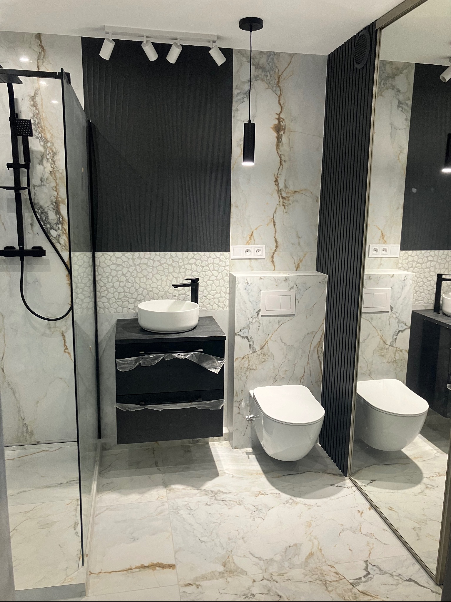 Nowoczesna łazienka z marmurową podłogą i ścianami, czarną szafką pod umywalką i kabiną prysznicową ze szklaną ścianą. Minimalistyczne oświetlenie.