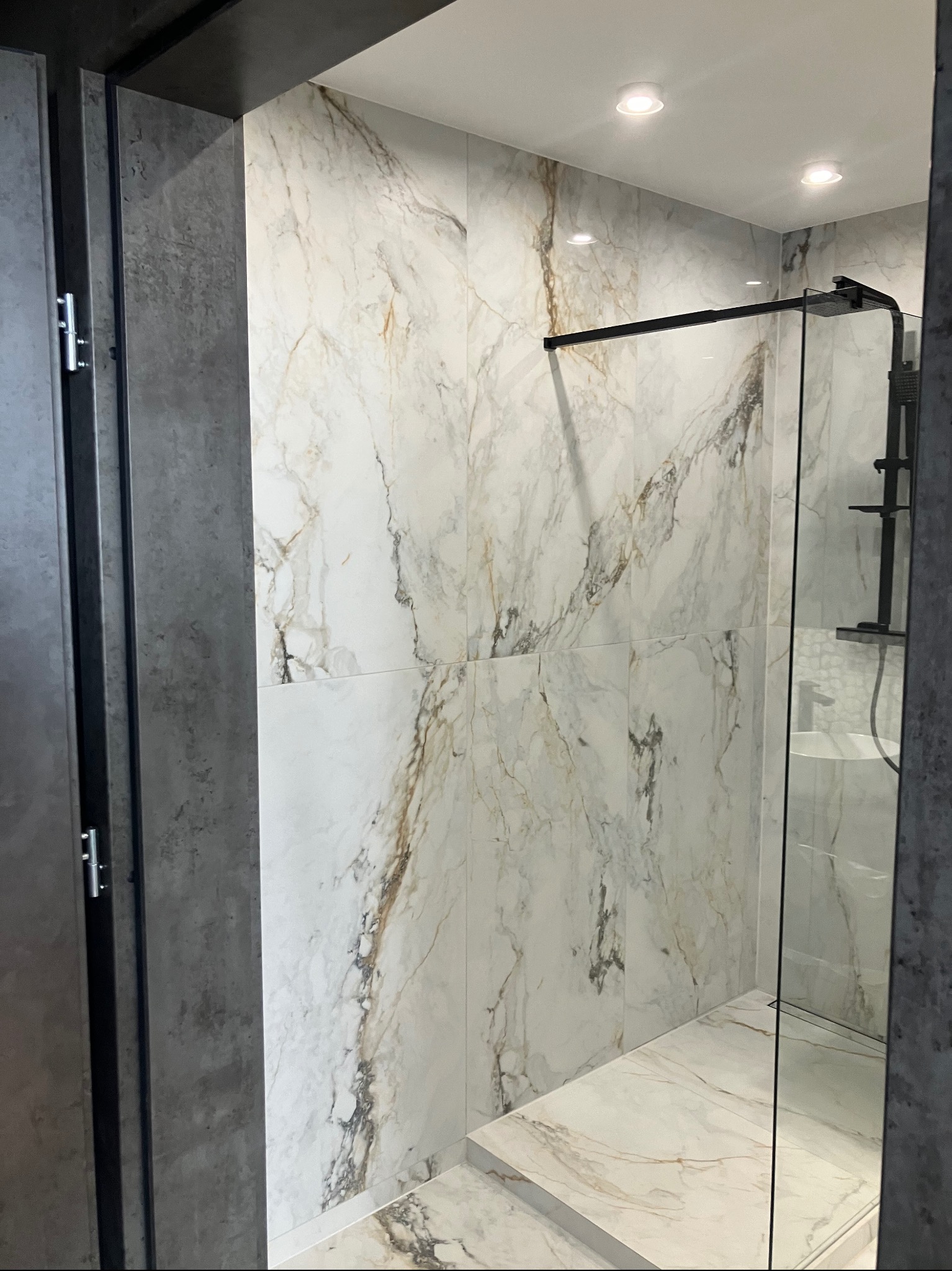 Nowoczesna kabina prysznicowa z marmurowymi płytkami w odcieniach bieli i złota, czarna armatura, szklana ściana. Minimalistyczny design, eleganckie wykończenie.