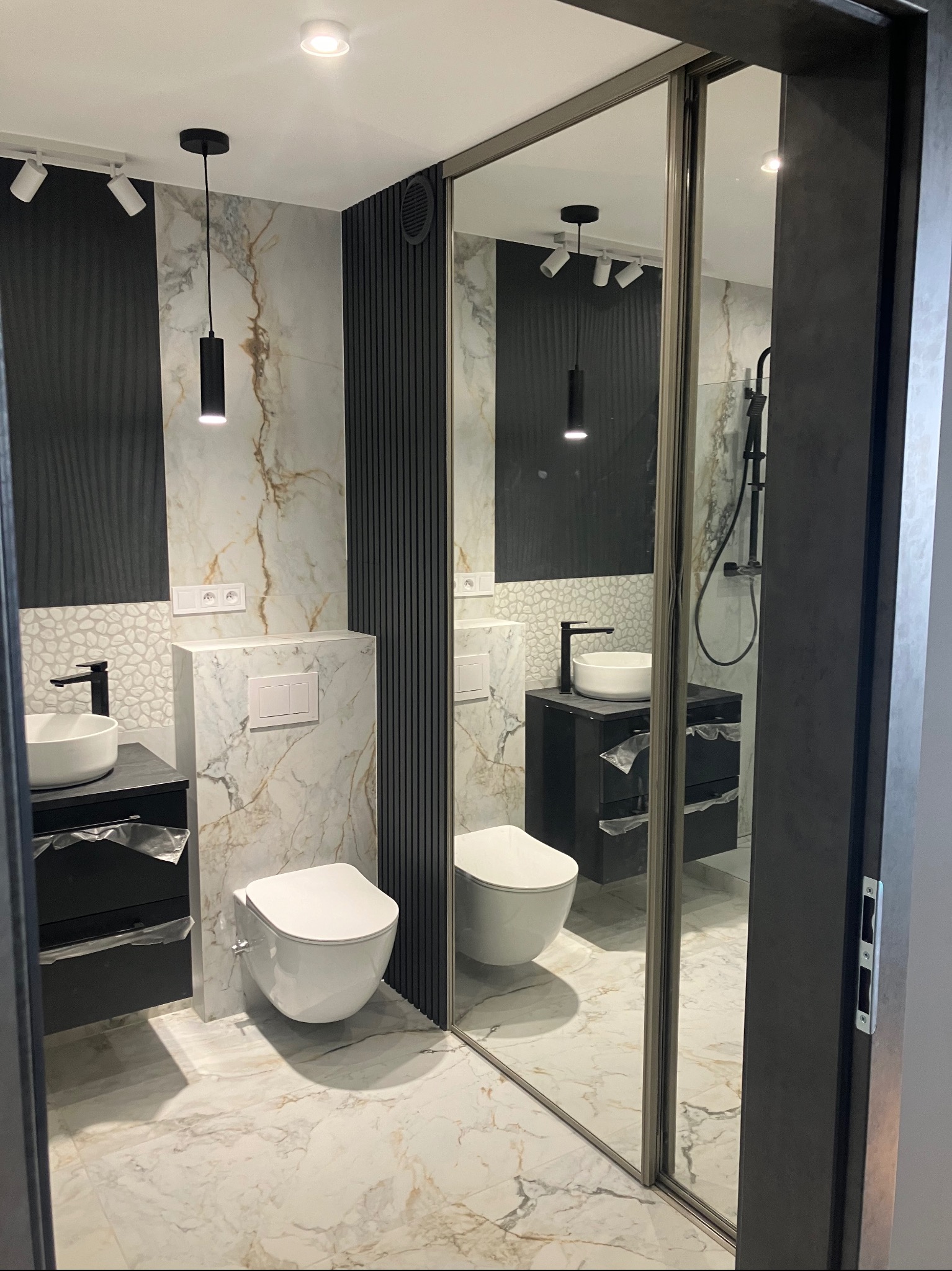 Nowoczesna łazienka z marmurowymi płytkami, czarną armaturą i oświetleniem punktowym. Umywalka nablatowa na szafce, wisząca toaleta i lustro z przesuwnymi drzwiami.