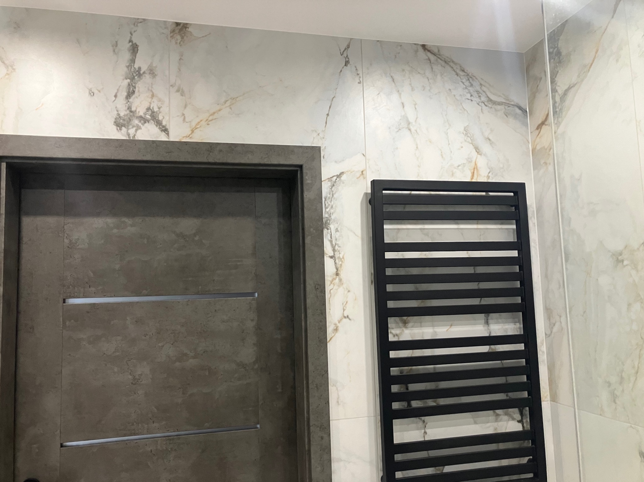 Wykończona łazienka z marmurowymi płytkami, ciemnymi drzwiami i czarnym grzejnikiem drabinkowym. Nowoczesny design z dbałością o detale.