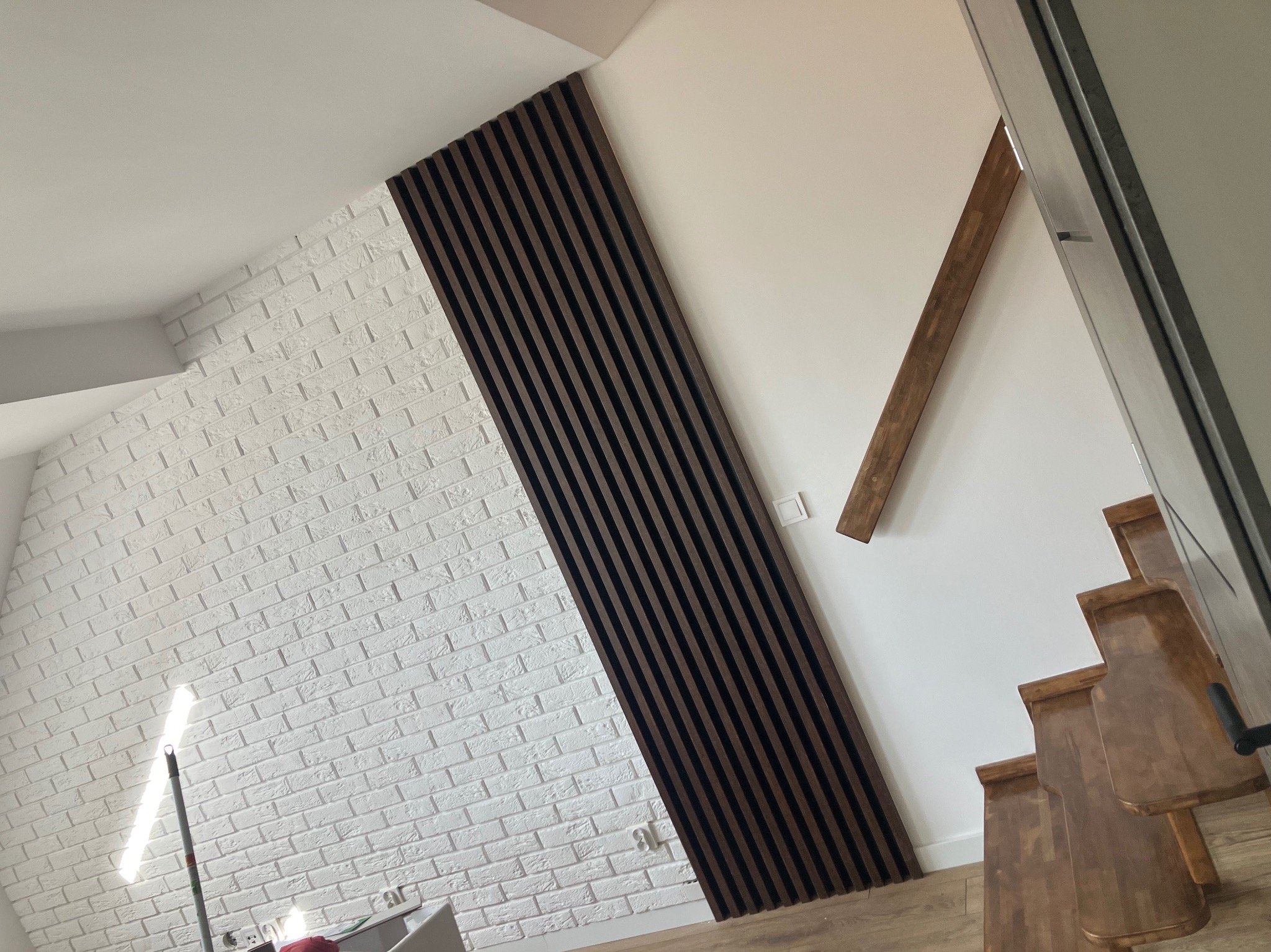 Wykończenie wnętrza w Wałbrzychu: biała cegła na ścianie zestawiona z dekoracyjnymi panelami lamelowymi w kolorze ciemnego drewna, widok z dołu.