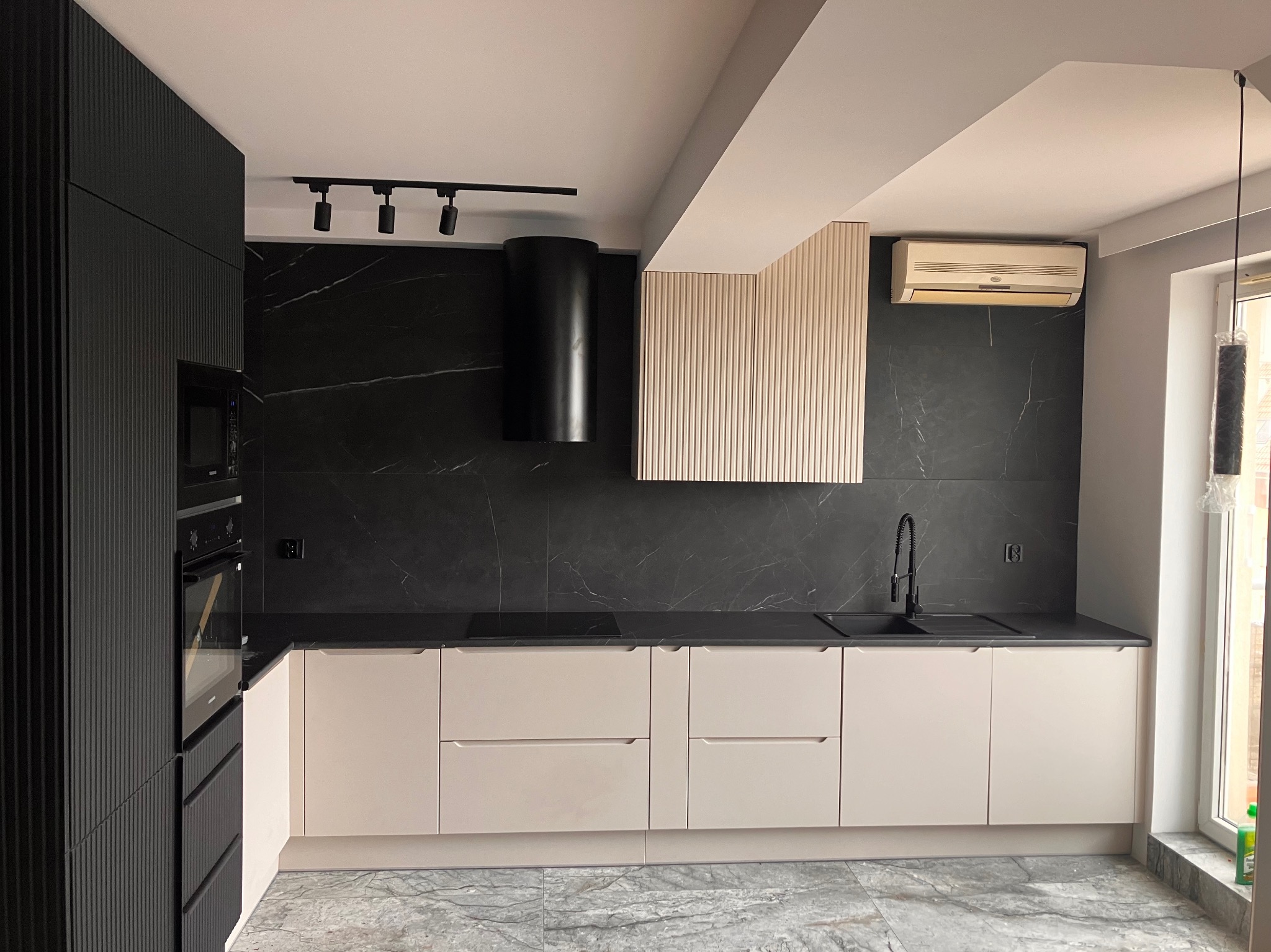 Nowoczesna kuchnia z czarnym marmurem na ścianie, beżowymi szafkami i czarnymi akcentami: okap, bateria i oświetlenie. Minimalistyczny design z klimatyzacją.
