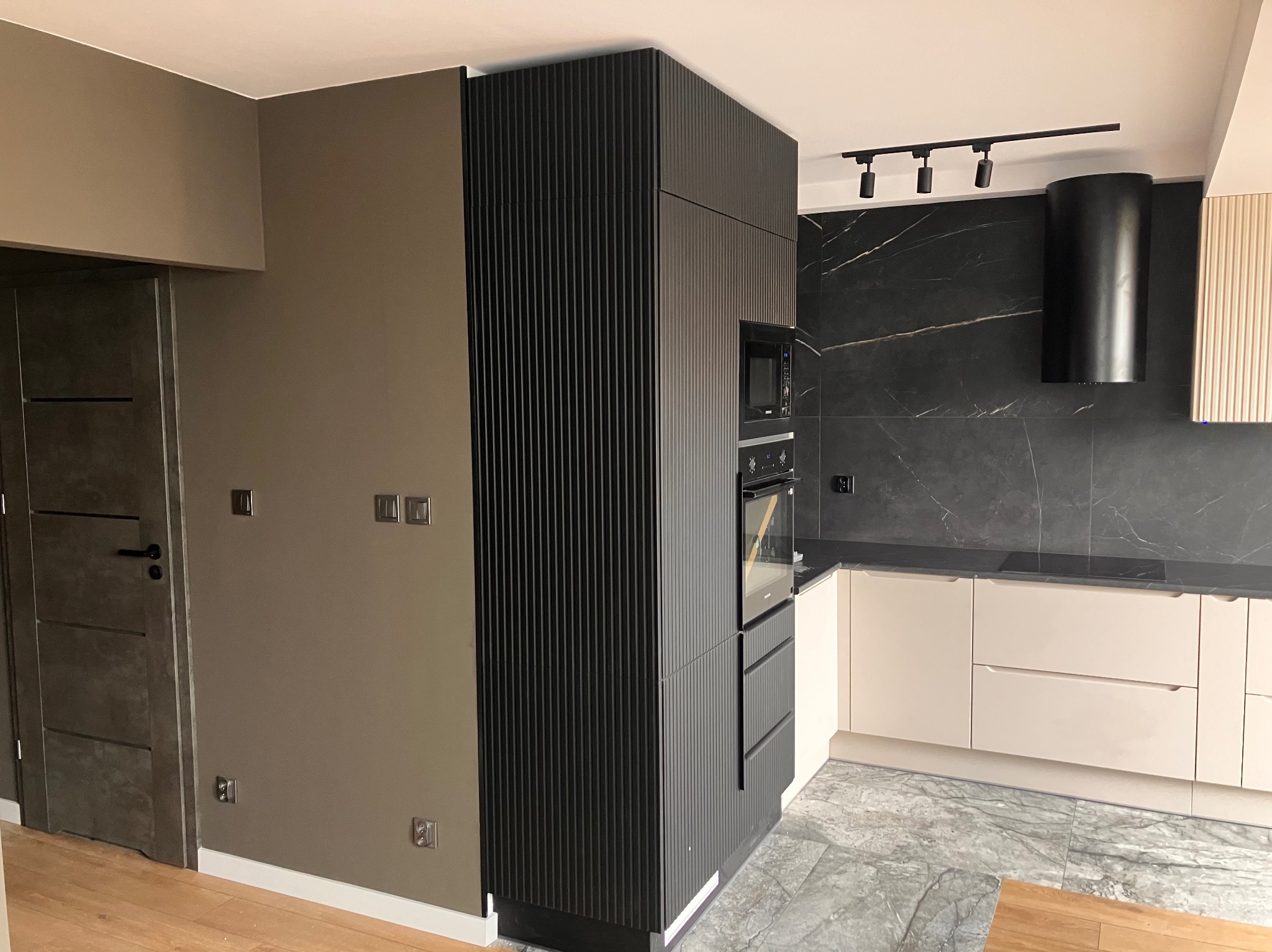 Nowoczesna kuchnia z czarnymi, ryflowanymi szafkami, marmurowym blatem i szarą podłogą. Widoczne oświetlenie punktowe i minimalistyczny okap.
