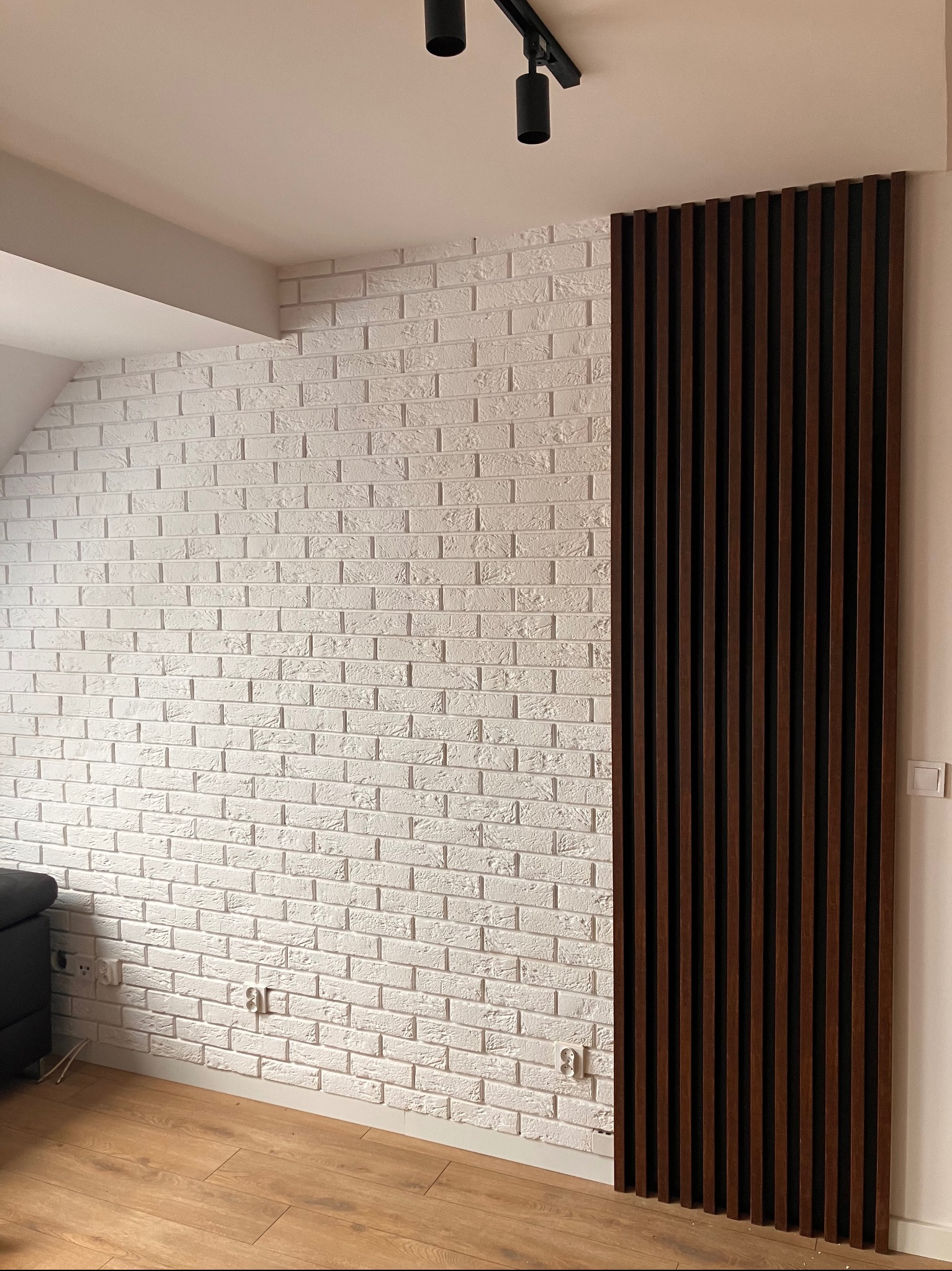 Biała ściana z cegły dekoracyjnej sąsiadująca z panelem lamelowym w odcieniach brązu. Widoczne gniazdka elektryczne i oświetlenie sufitowe.