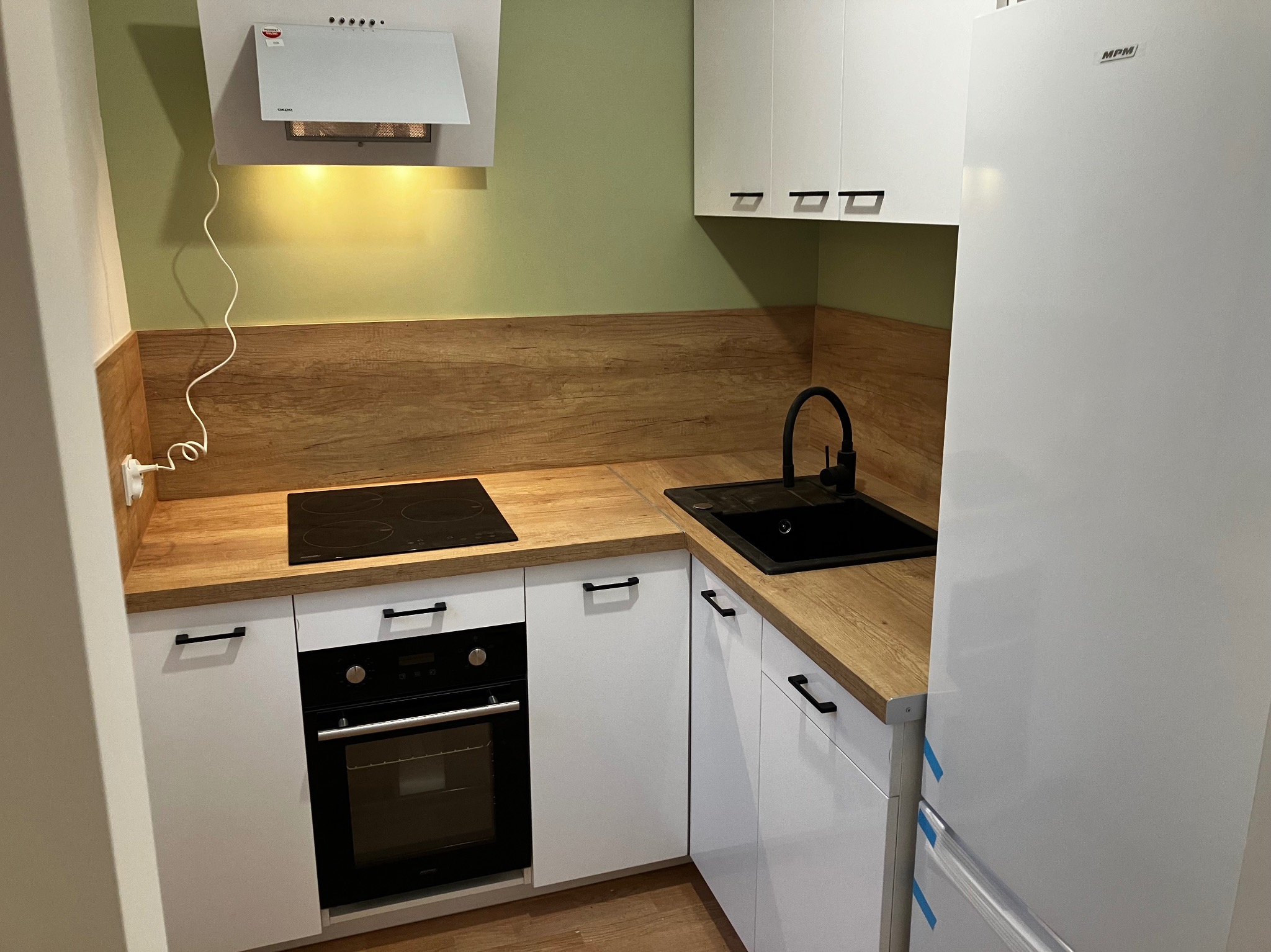 Nowoczesna kuchnia z białymi szafkami, drewnianym blatem i czarną armaturą. Widoczny piekarnik, płyta indukcyjna i lodówka MPM. Ściana w kolorze oliwkowym.