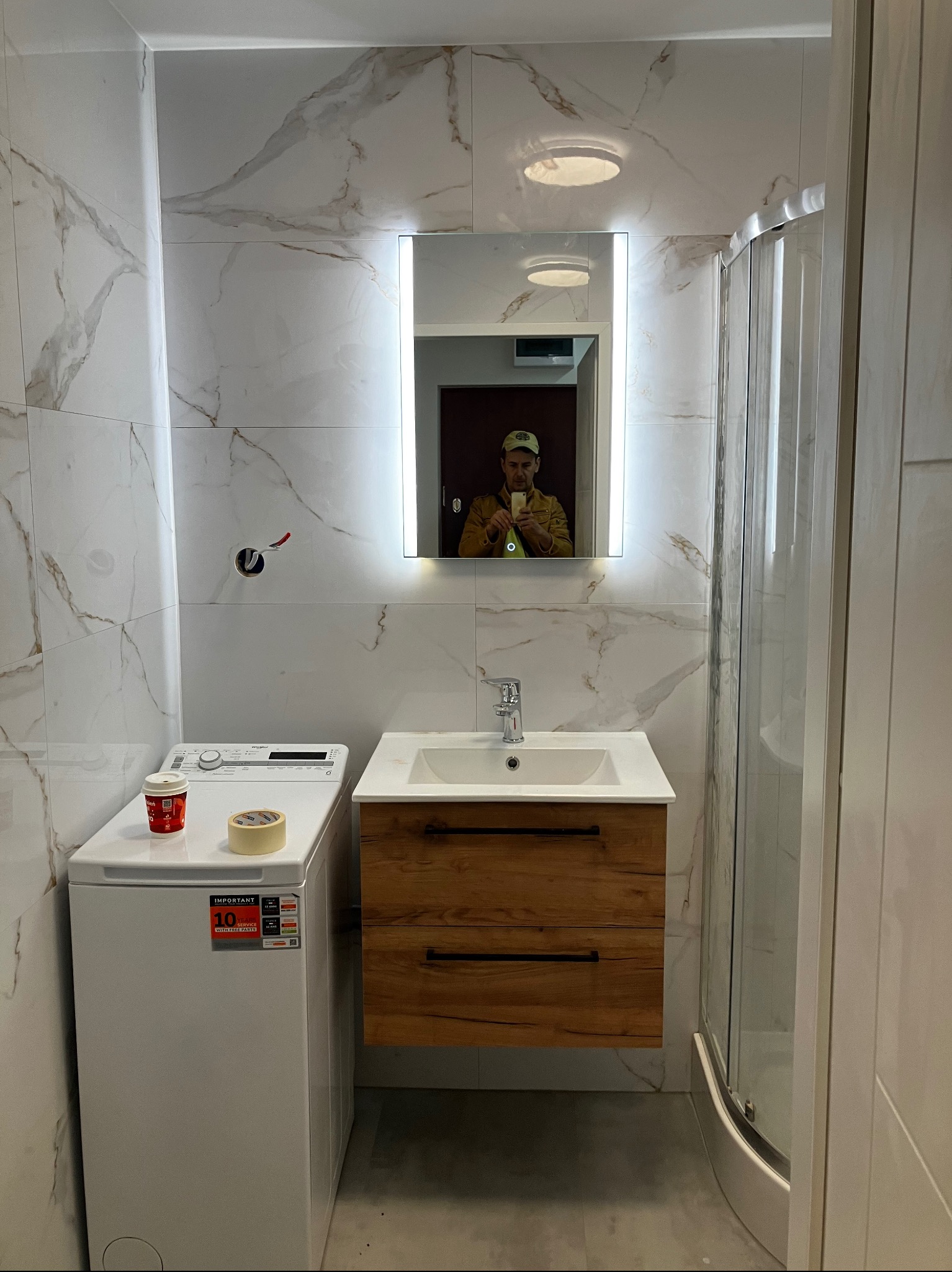 Wykończona łazienka z kabiną prysznicową, umywalką z szafką z drewnianym frontem i pralką. Na blacie kubek i taśma malarska. Ściany wyłożone płytkami imitującymi marmur.