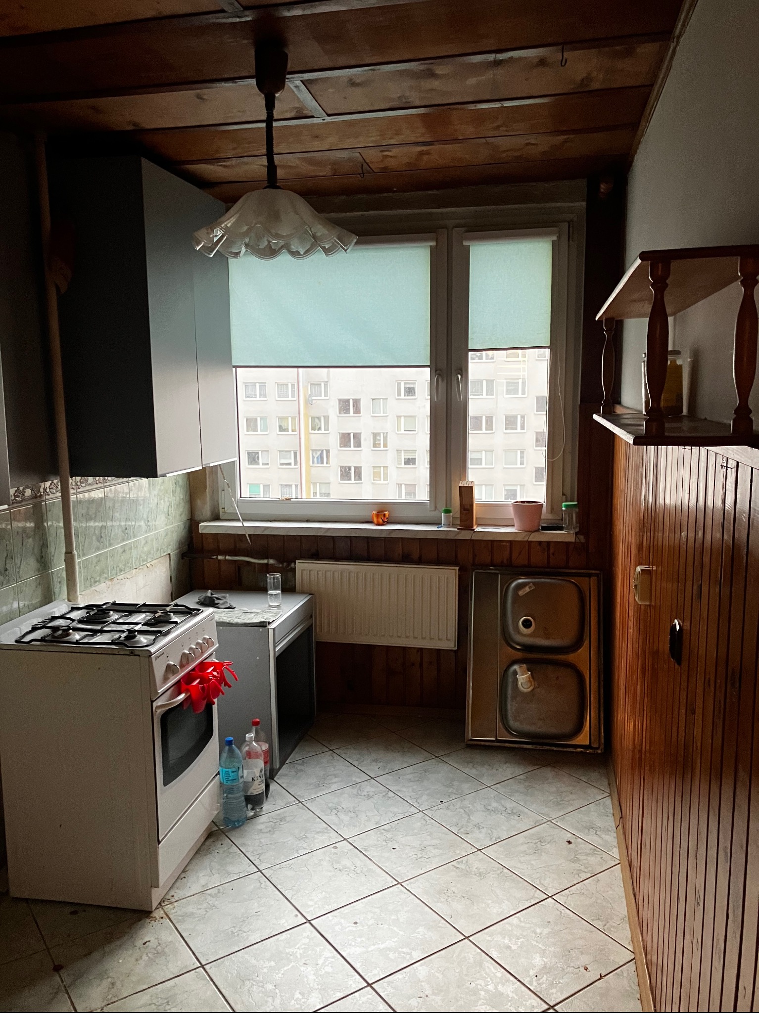 Kadr z kuchni przed remontem: stara kuchenka, okno z roletą, drewniana boazeria i płytki podłogowe. Widoczne ślady użytkowania i zużycia.