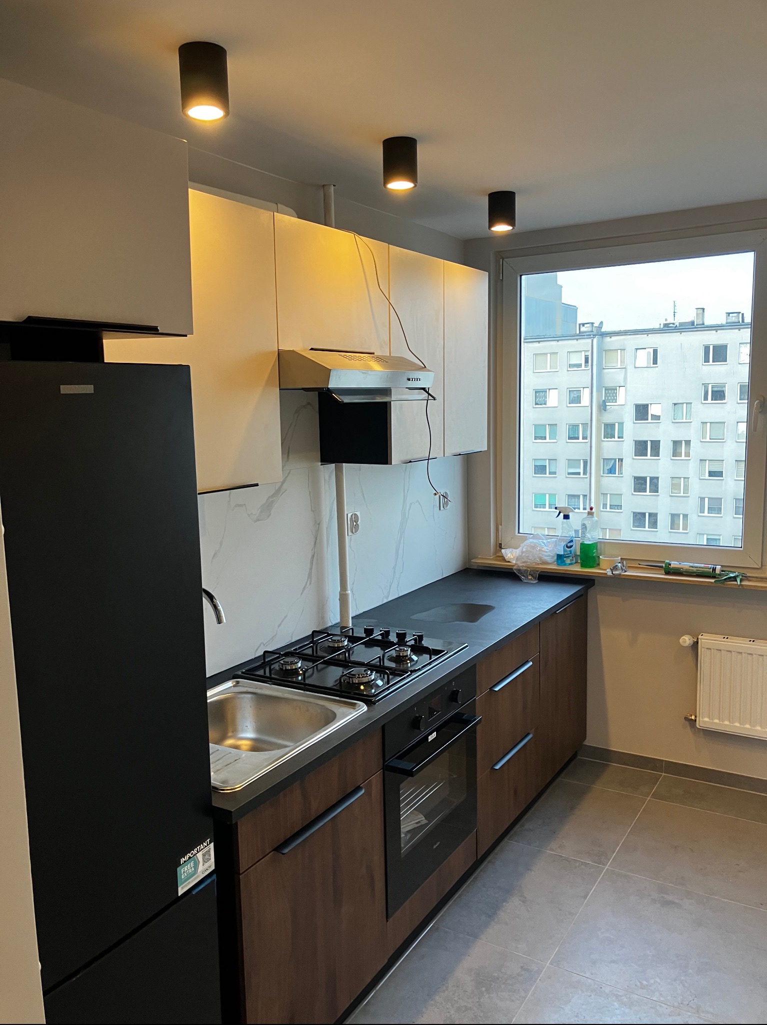 Nowoczesna kuchnia z czarną lodówką, drewnianymi szafkami i marmurowym backsplashiem. Widok na budynek przez okno. Minimalistyczne oświetlenie sufitowe.