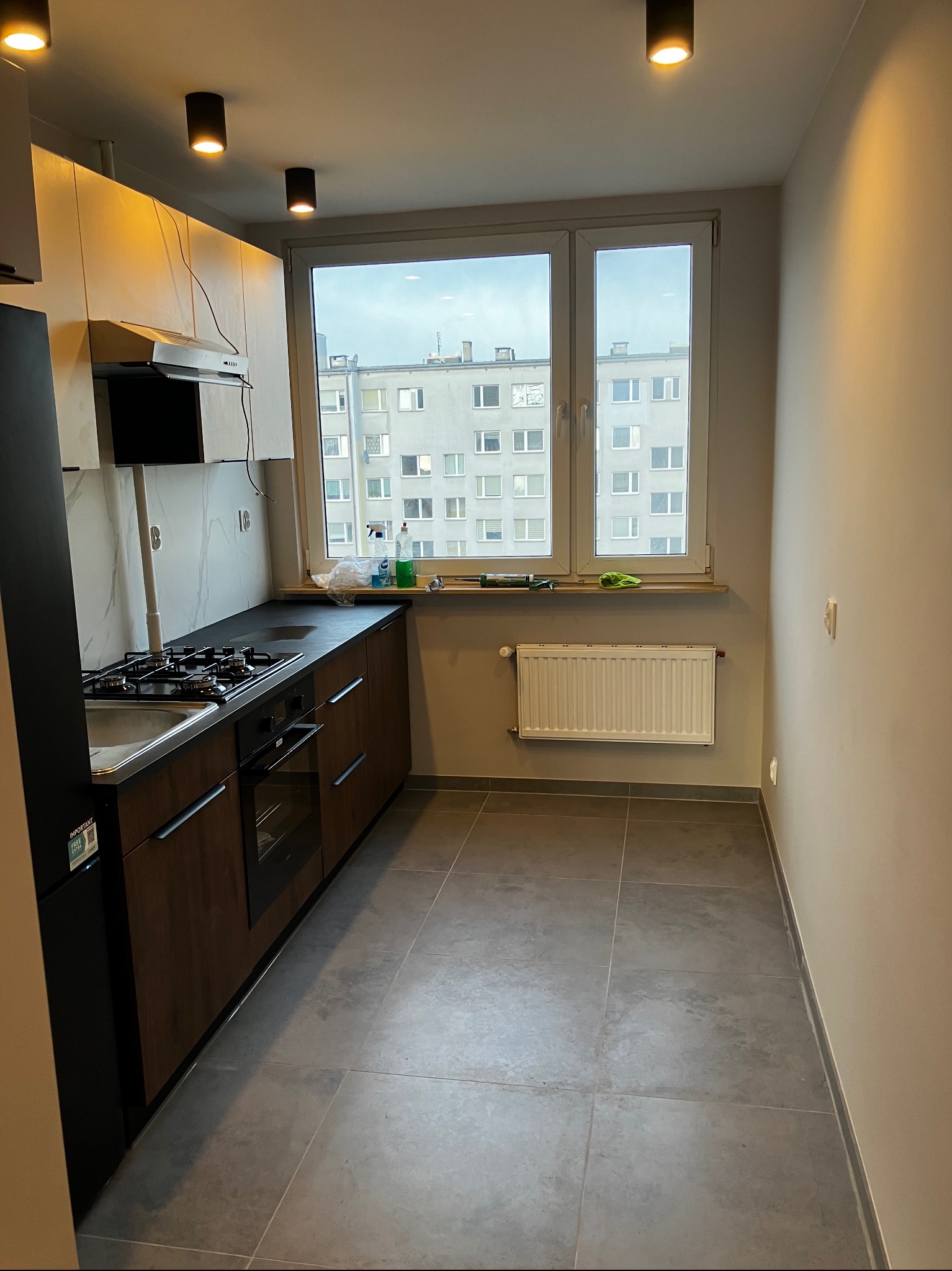 Nowoczesna kuchnia z ciemnymi szafkami i szarą podłogą. Okno z widokiem na budynki. Minimalistyczne oświetlenie sufitowe. Umywalka, płyta grzewcza i piekarnik w zabudowie.