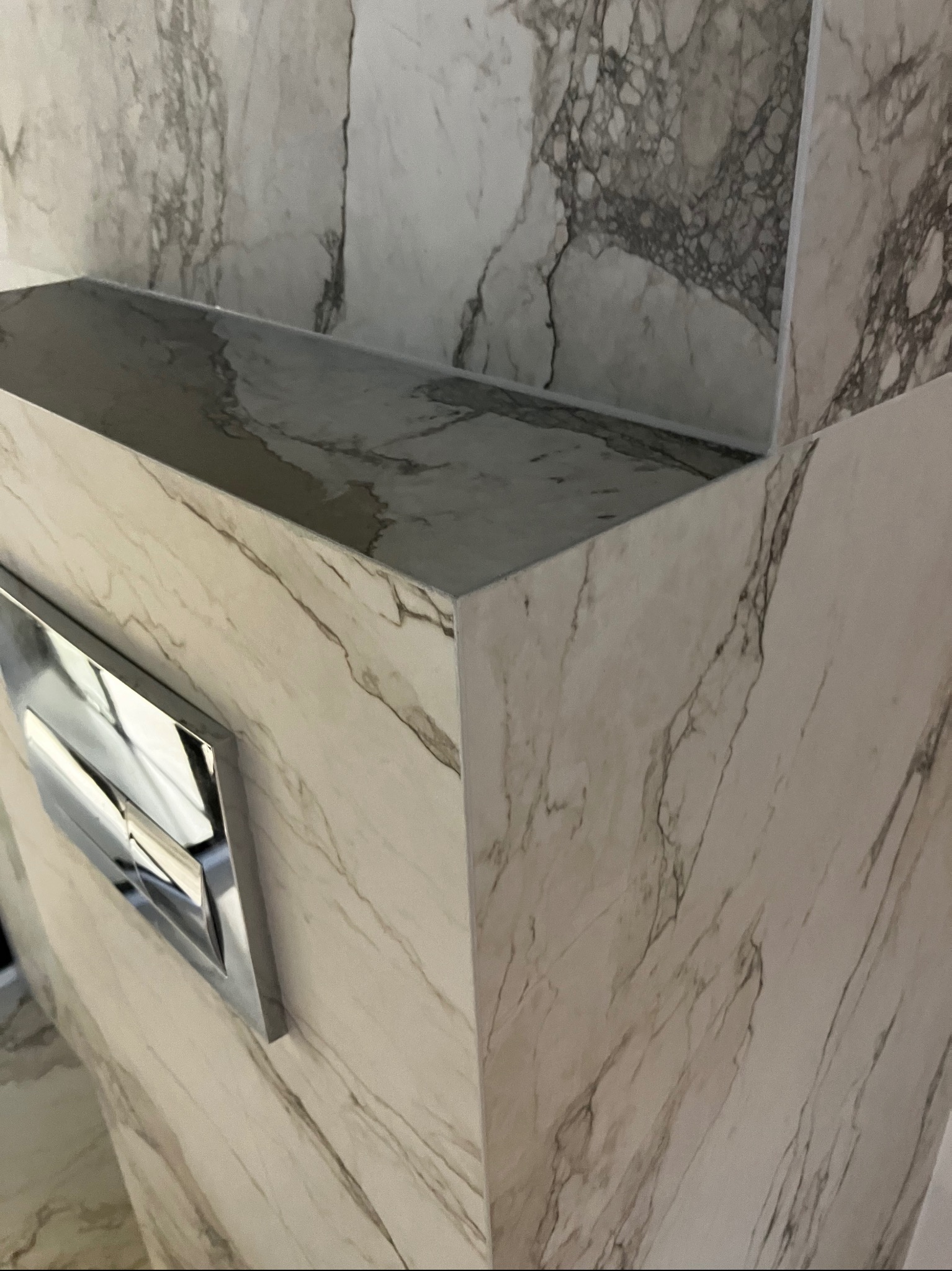 Wykończona toaleta w łazience z marmurowymi płytkami i chromowanym przyciskiem spłukiwania. Minimalistyczny design, eleganckie wykończenie.