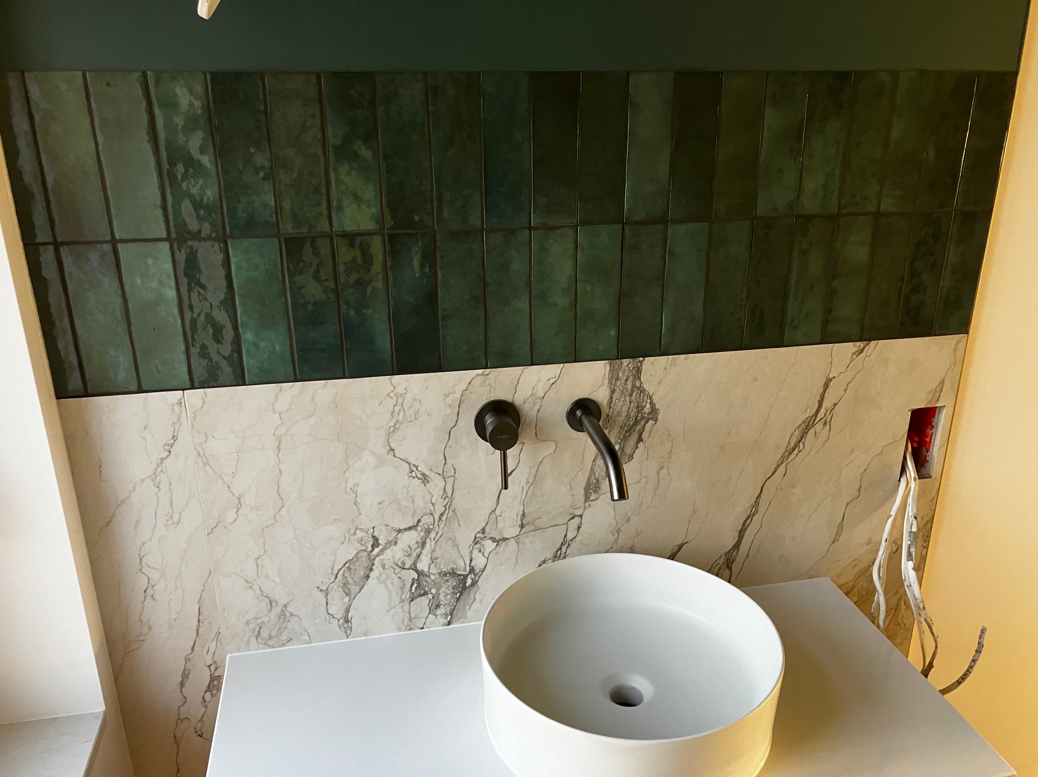 Nowoczesna łazienka: umywalka nablatowa, bateria w kolorze antracytu, ściana z płytek imitujących marmur i zielona glazura wertykalna. Widoczne przewody elektryczne.