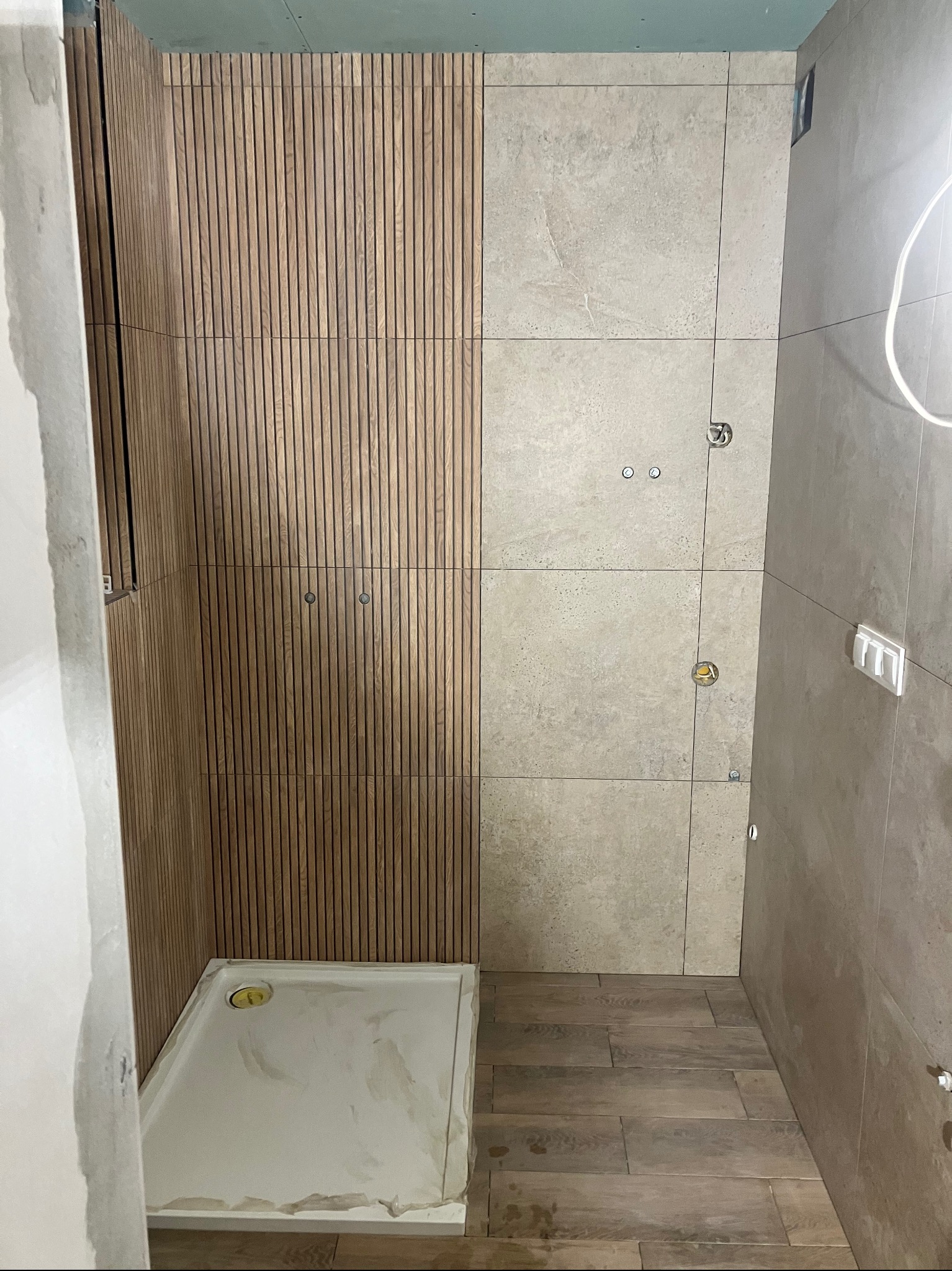 Wykończona łazienka z drewnianą ścianą z lameli i beżowymi płytkami. Widoczny brodzik i fragment podłogi z płytek imitujących drewno. Przygotowane punkty instalacji.