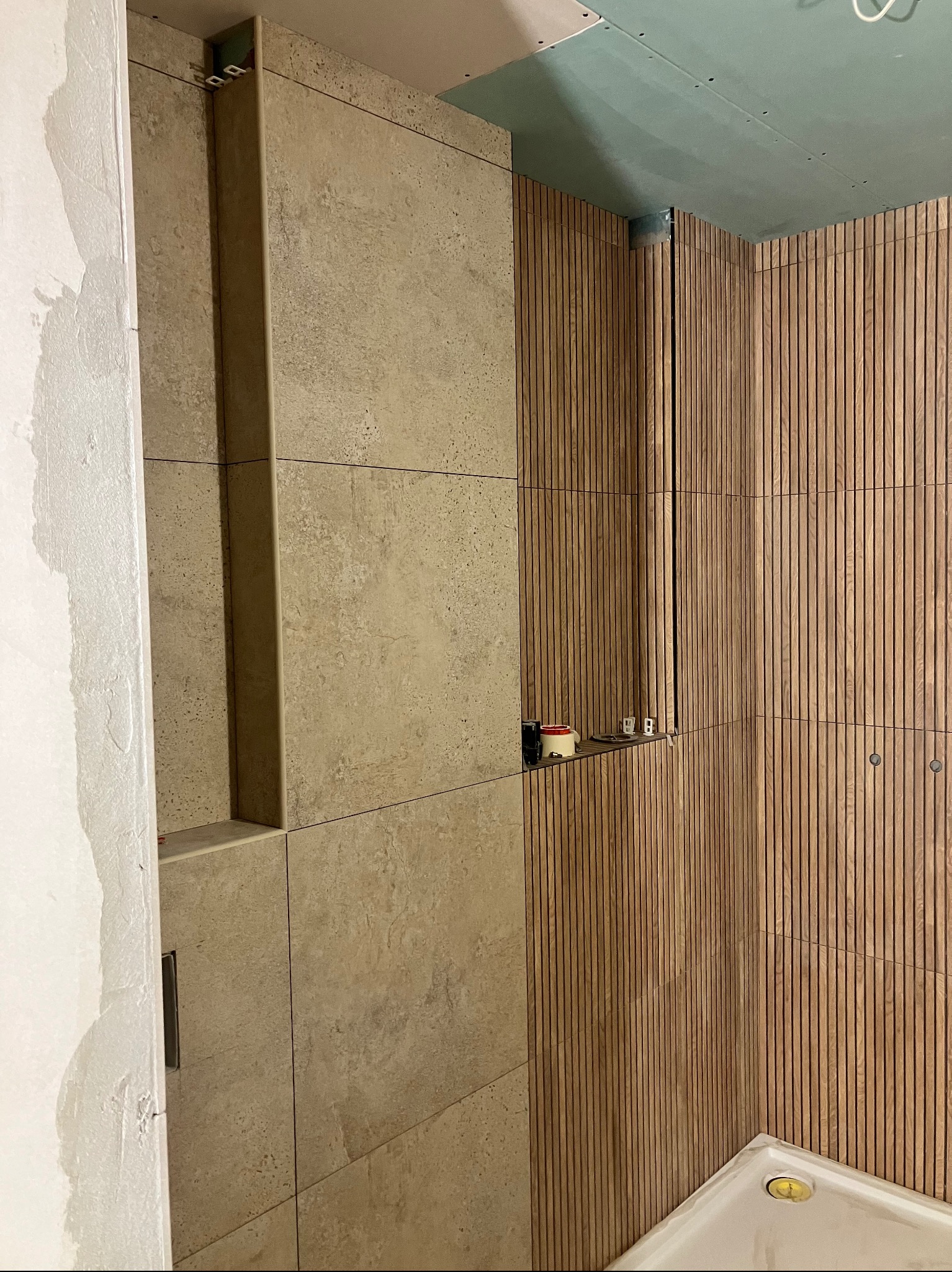 Wykończona ściana łazienki z beżowych płytek i drewnianych paneli. Widoczne gniazdka elektryczne i fragment sufitu z płyt karton-gips. Detal wykończenia wnętrza.