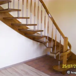 Jasne, drewniane schody z metalowymi wspornikami balustrady, widok z dołu, z datą i adresem strony internetowej w prawym dolnym rogu.