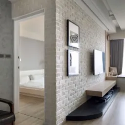 Fragment wnętrza: ściana z cegły pomalowanej na biało, z dwoma obrazami w czarnych ramkach i telewizorem, drewniana podłoga imitująca panele, widok na sypialnię przez otwarte drzwi.