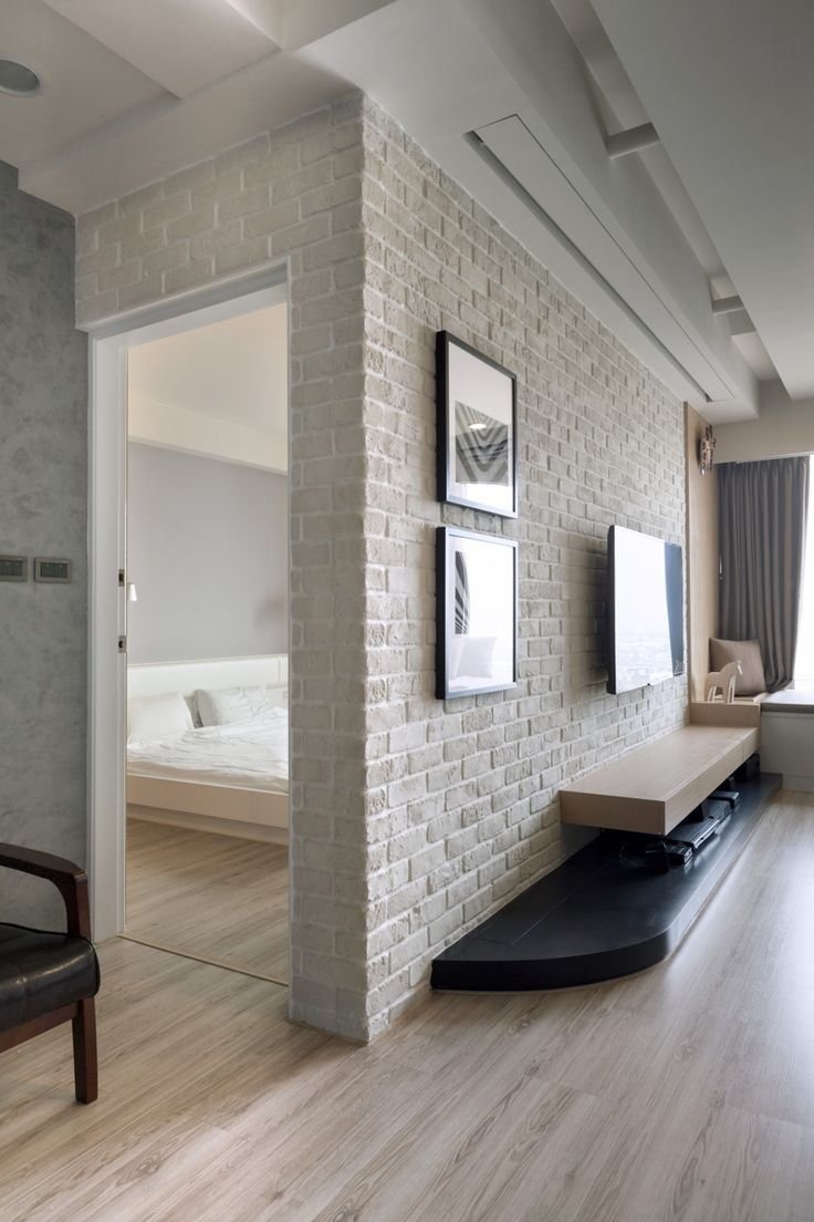 Fragment wnętrza: ściana z cegły pomalowanej na biało, z dwoma obrazami w czarnych ramkach i telewizorem, drewniana podłoga imitująca panele, widok na sypialnię przez otwarte drzwi.