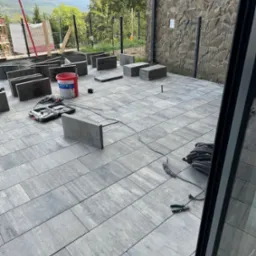 Montaż szarej kostki brukowej na tarasie z widokiem na góry, widoczny poziomica, młotek i wiadro z zaprawą.