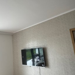 Foxybud - Telewizor zamontowany na ścianie z tapetą w odcieniach szarości, widoczne gniazdka elektryczne i czujnik dymu na suficie.