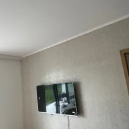 Telewizor zamontowany na ścianie z tapetą w odcieniach szarości, widoczne gniazdka elektryczne i czujnik dymu na suficie.