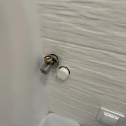 Szary zawór kątowy z mosiężną końcówką zamontowany na ścianie z białych płytek o falistej strukturze, obok otworu z wystającą białą rurką, w tle fragment białego pojemnika.