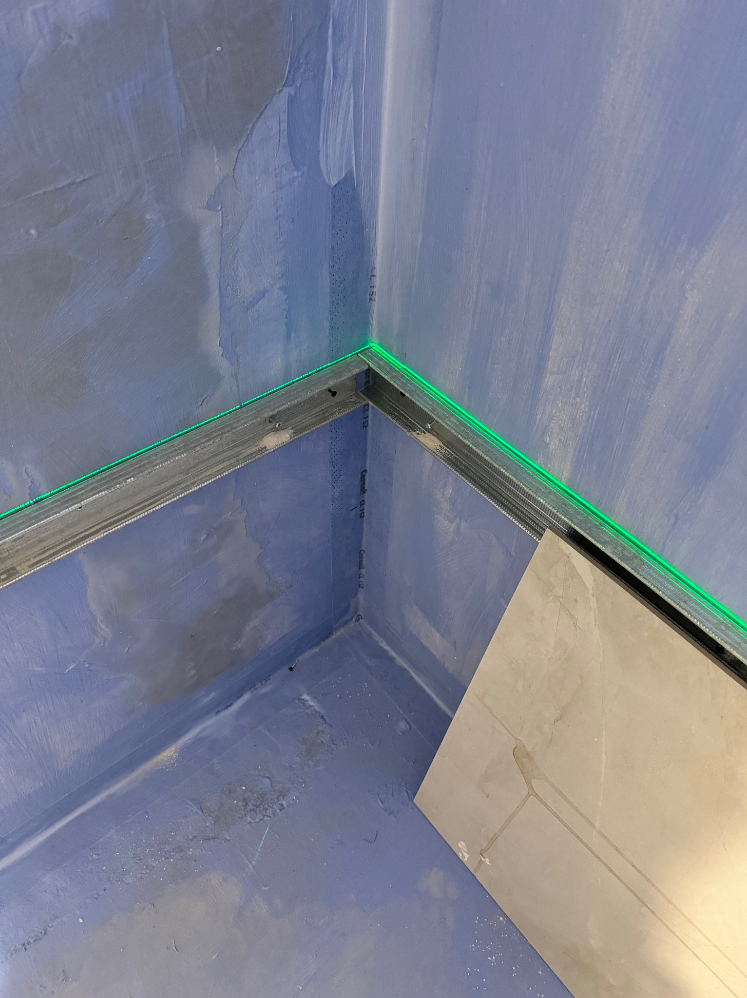 Przygotowanie do układania płytek w rogu pomieszczenia, widoczne profile przyścienne z zielonym laserem poziomującym i fragment płytki podłogowej.