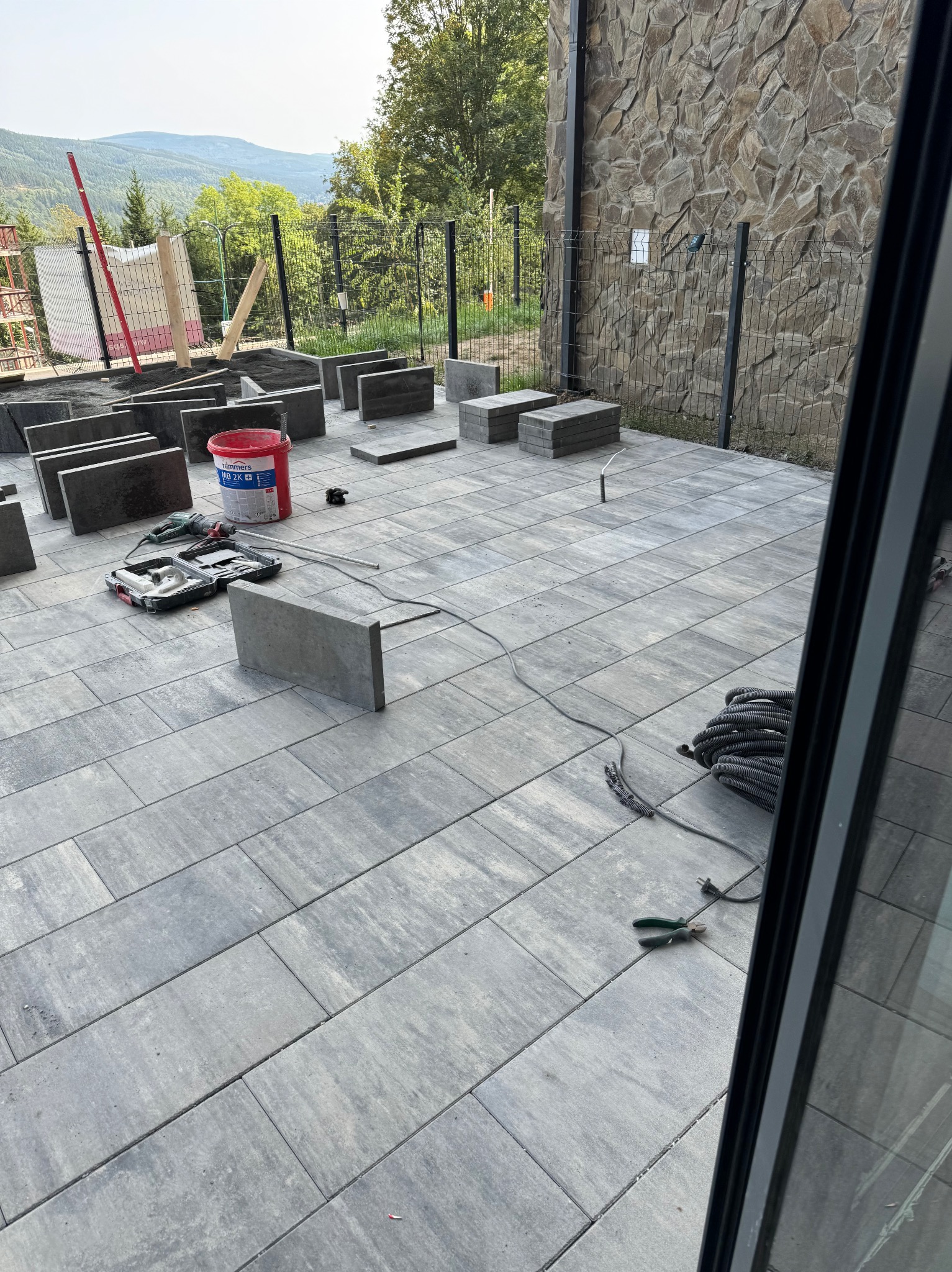 Montaż szarej kostki brukowej na tarasie z widokiem na góry, widoczny poziomica, młotek i wiadro z zaprawą.