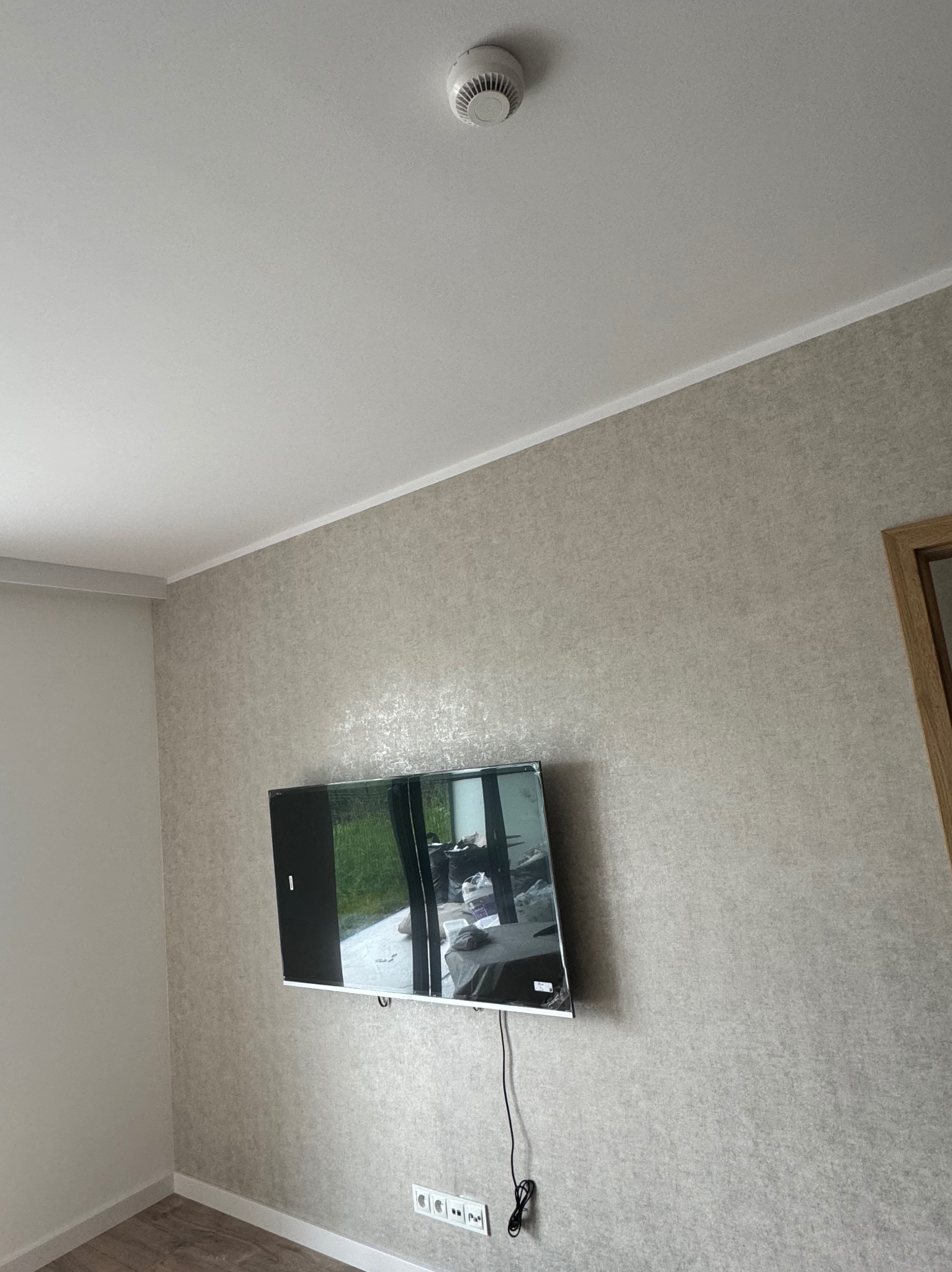 Telewizor zamontowany na ścianie z tapetą w odcieniach szarości, widoczne gniazdka elektryczne i czujnik dymu na suficie.