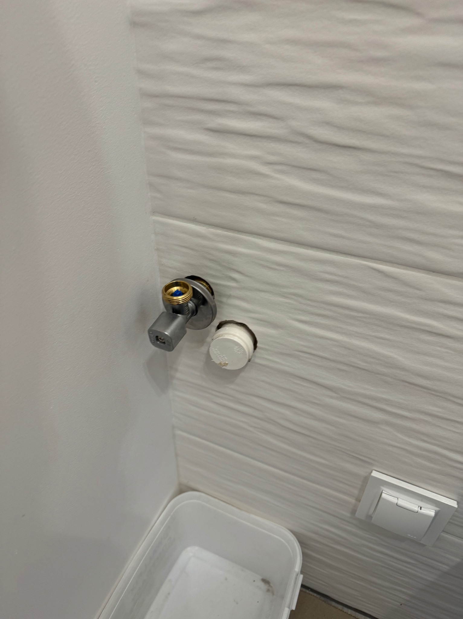 Szary zawór kątowy z mosiężną końcówką zamontowany na ścianie z białych płytek o falistej strukturze, obok otworu z wystającą białą rurką, w tle fragment białego pojemnika.