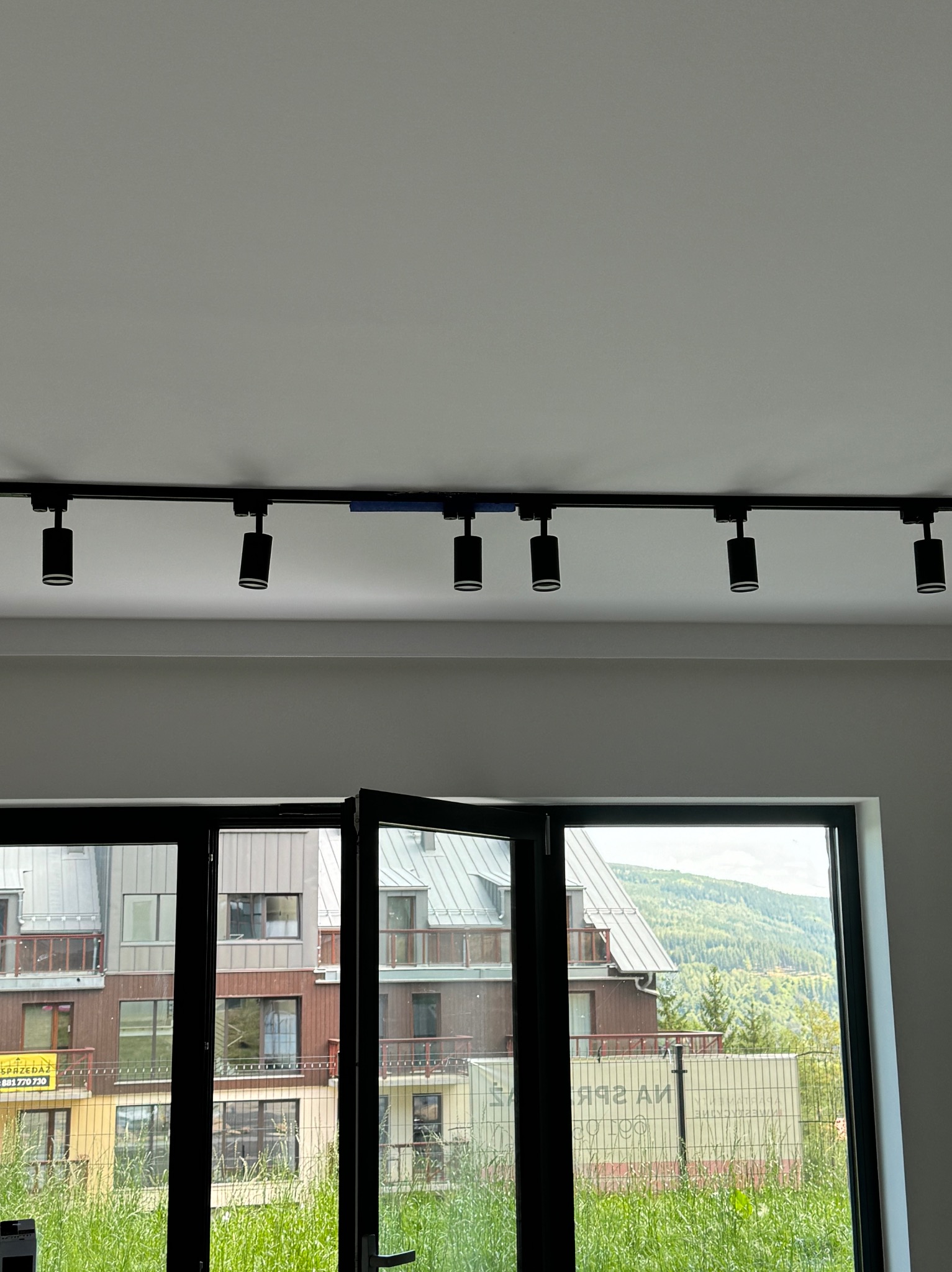 Sufit z zamontowanym czarnym systemem szynoprzewodów z reflektorami, widok przez otwarte okno na budynki mieszkalne i zieleń.